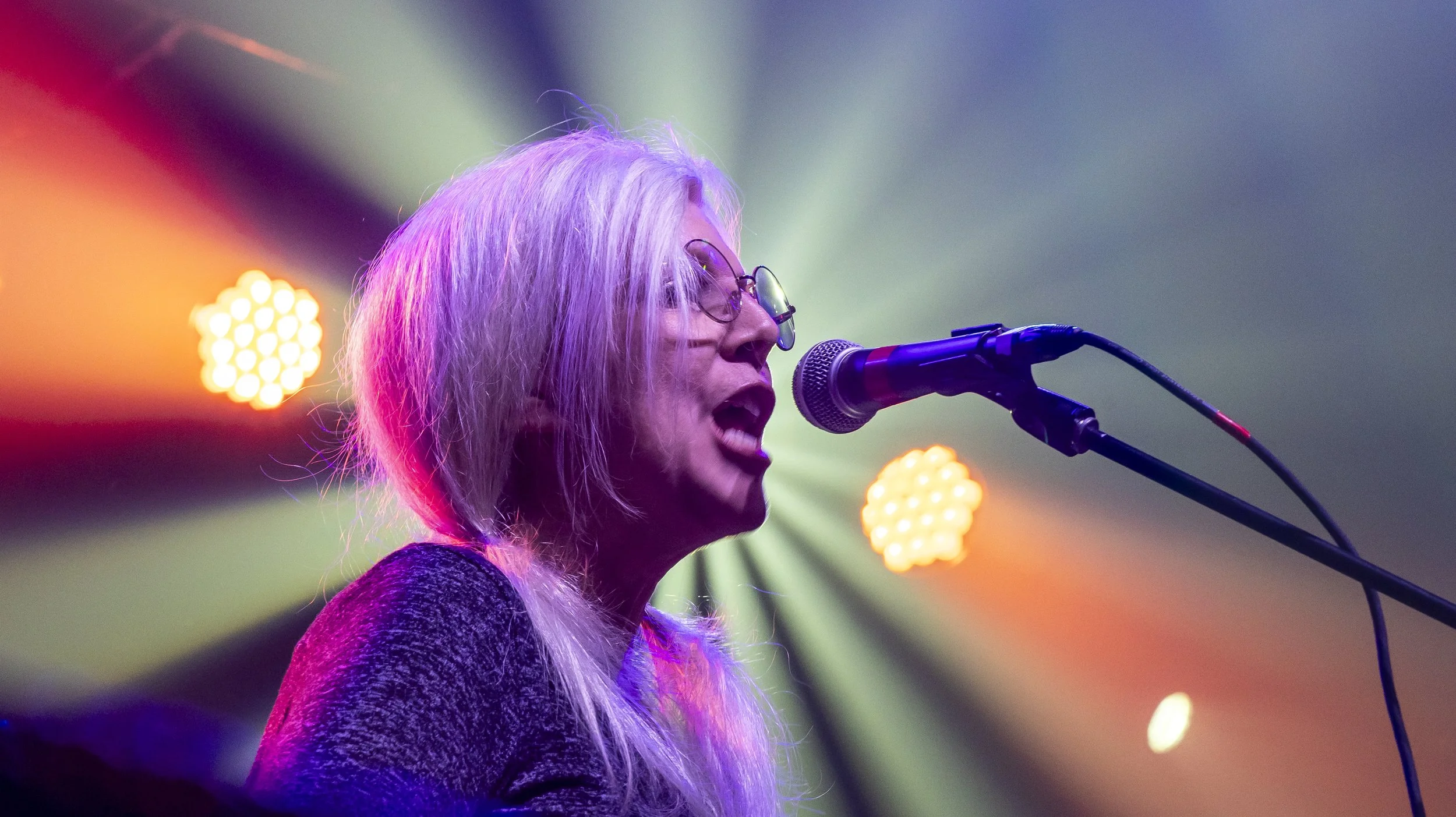 A woman with long blonde hair and glasses singing into a microphone on stage, lit by colorful stage lights.