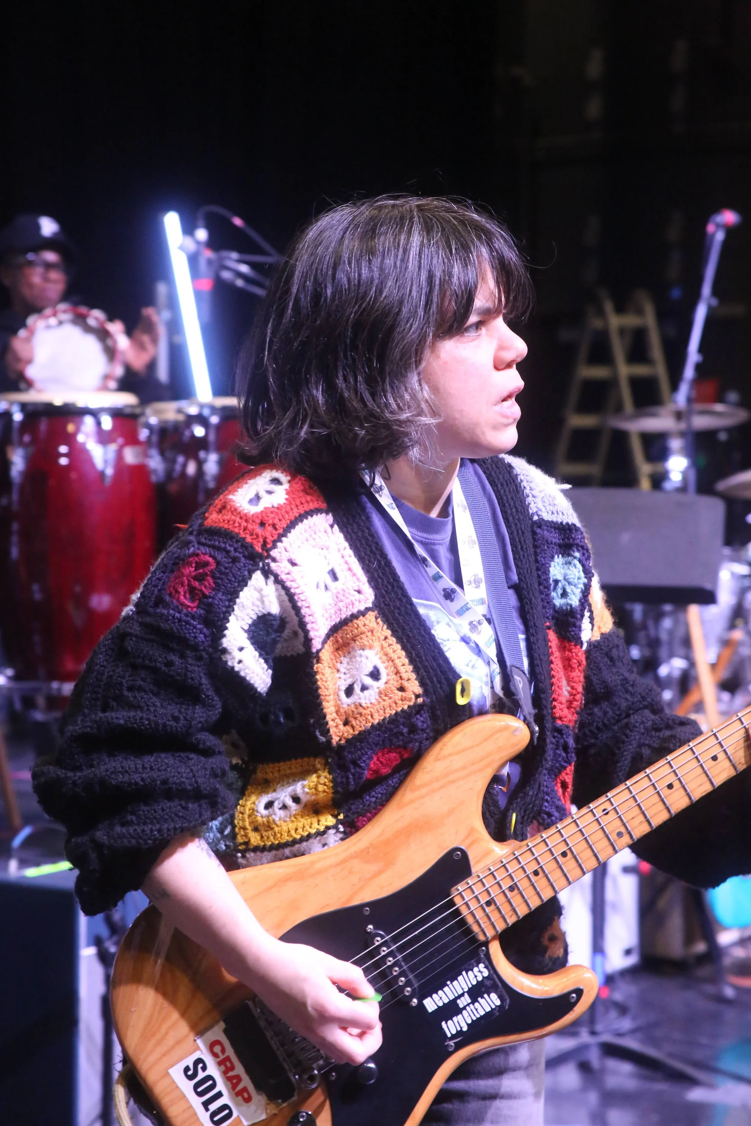 A person with shoulder-length dark hair playing an electric guitar, wearing a colorful crocheted sweater with patchwork designs. In the background, a drummer is visible, smiling, playing conga drums, with stage equipment and microphones around.