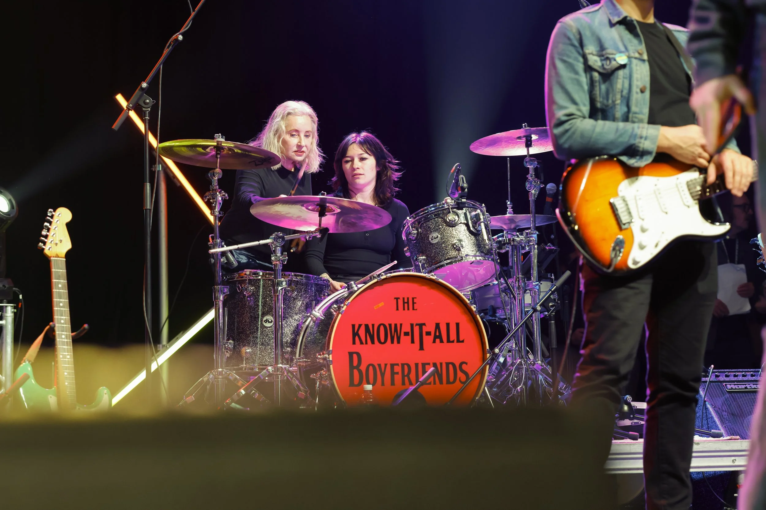 A band performing on stage with a drummer and guitarist, and a sign on the drum set reading 'THE KNOW-IT-ALL BOYFRIENDS'.