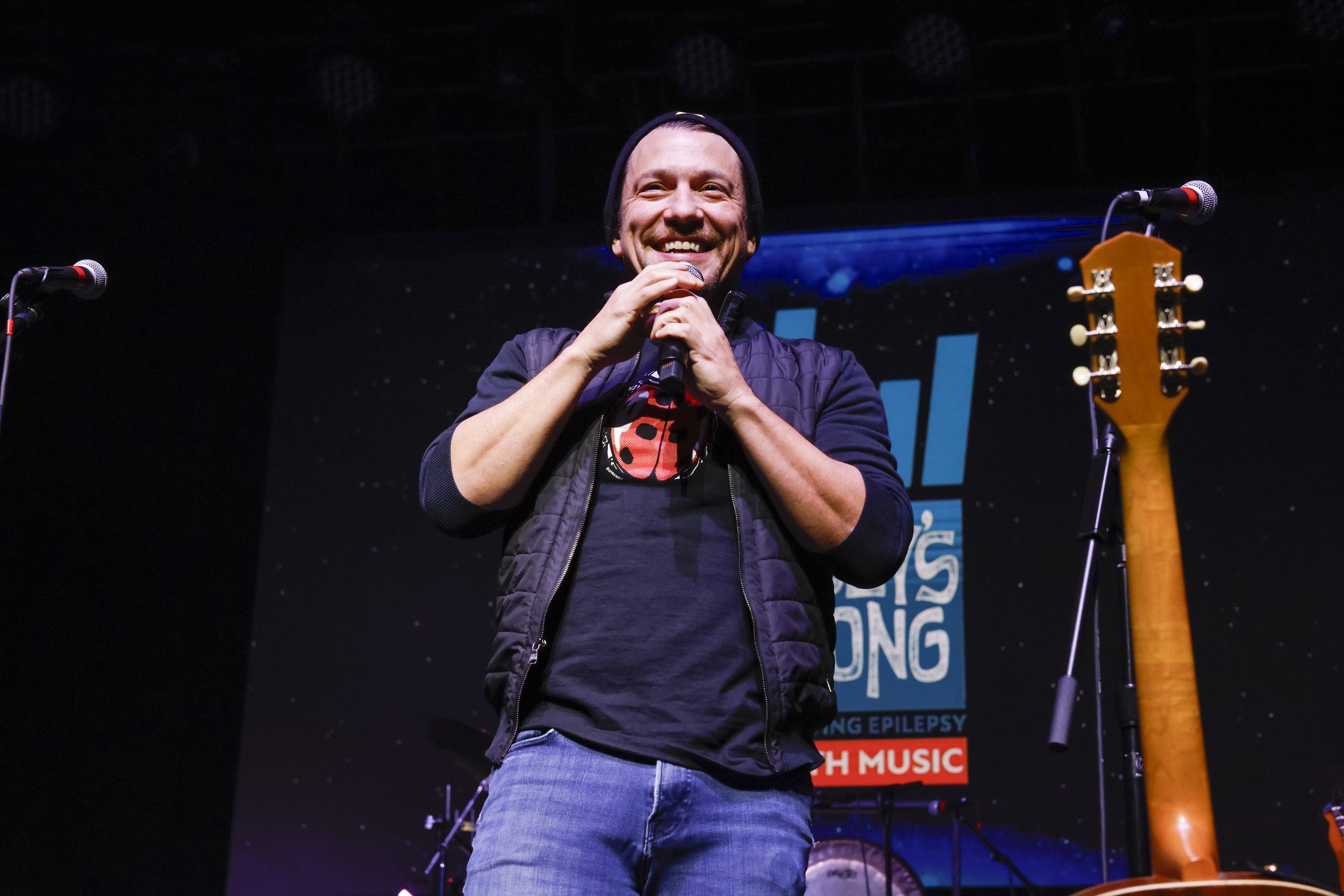 A man smiling and holding a microphone on a stage, wearing a black beanie, black jacket, and blue jeans, with two microphones on stands and a guitar nearby.