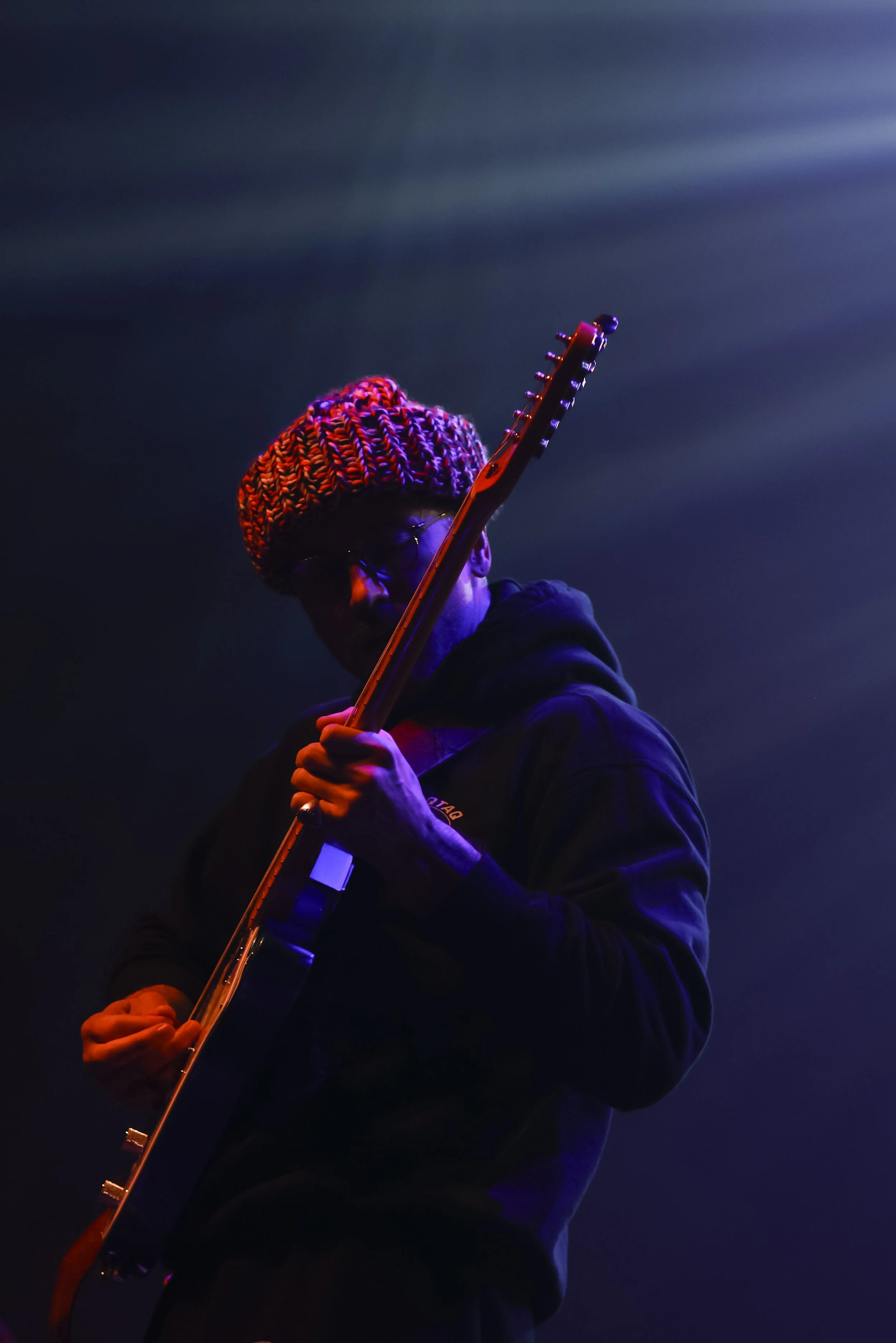 A person wearing a colorful knitted beanie playing an electric guitar with a dark background and purple lighting.