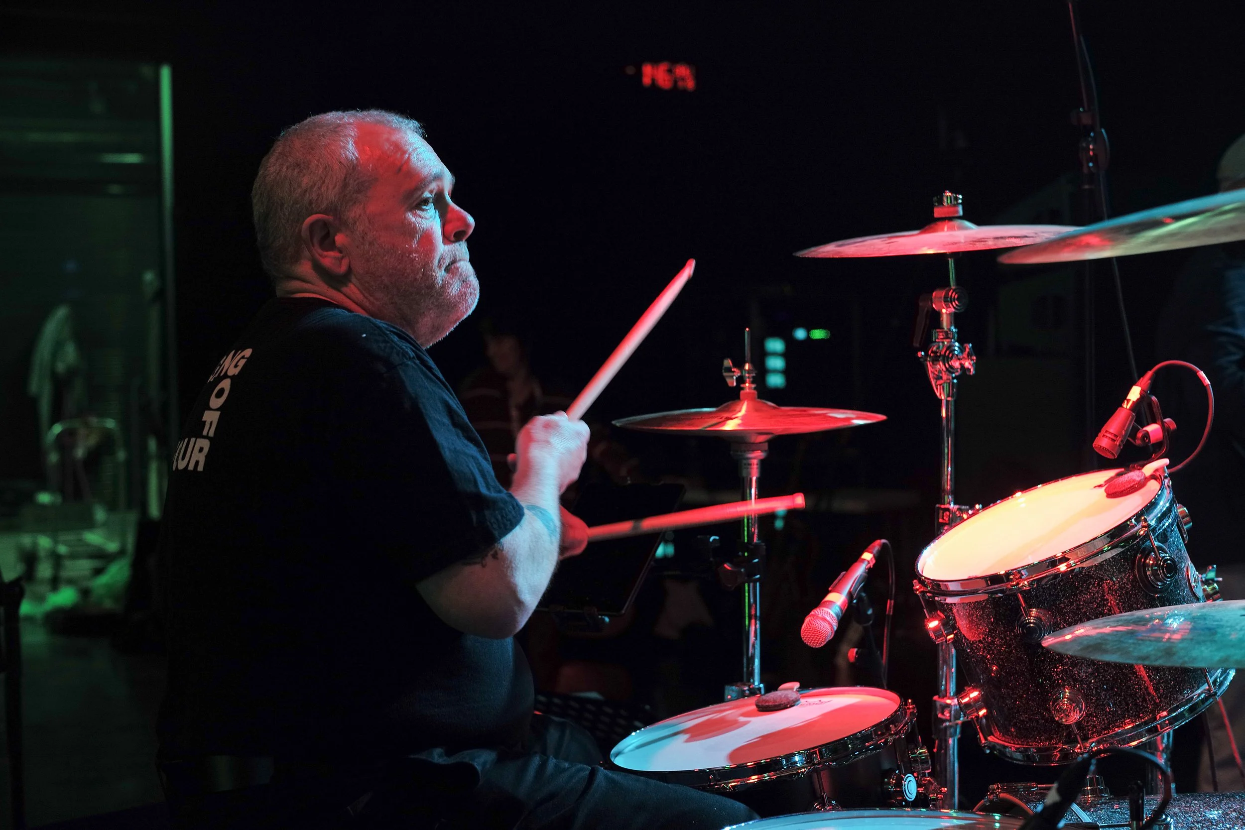 A man playing drums on stage with red and green lighting, focused and intense on his performance.
