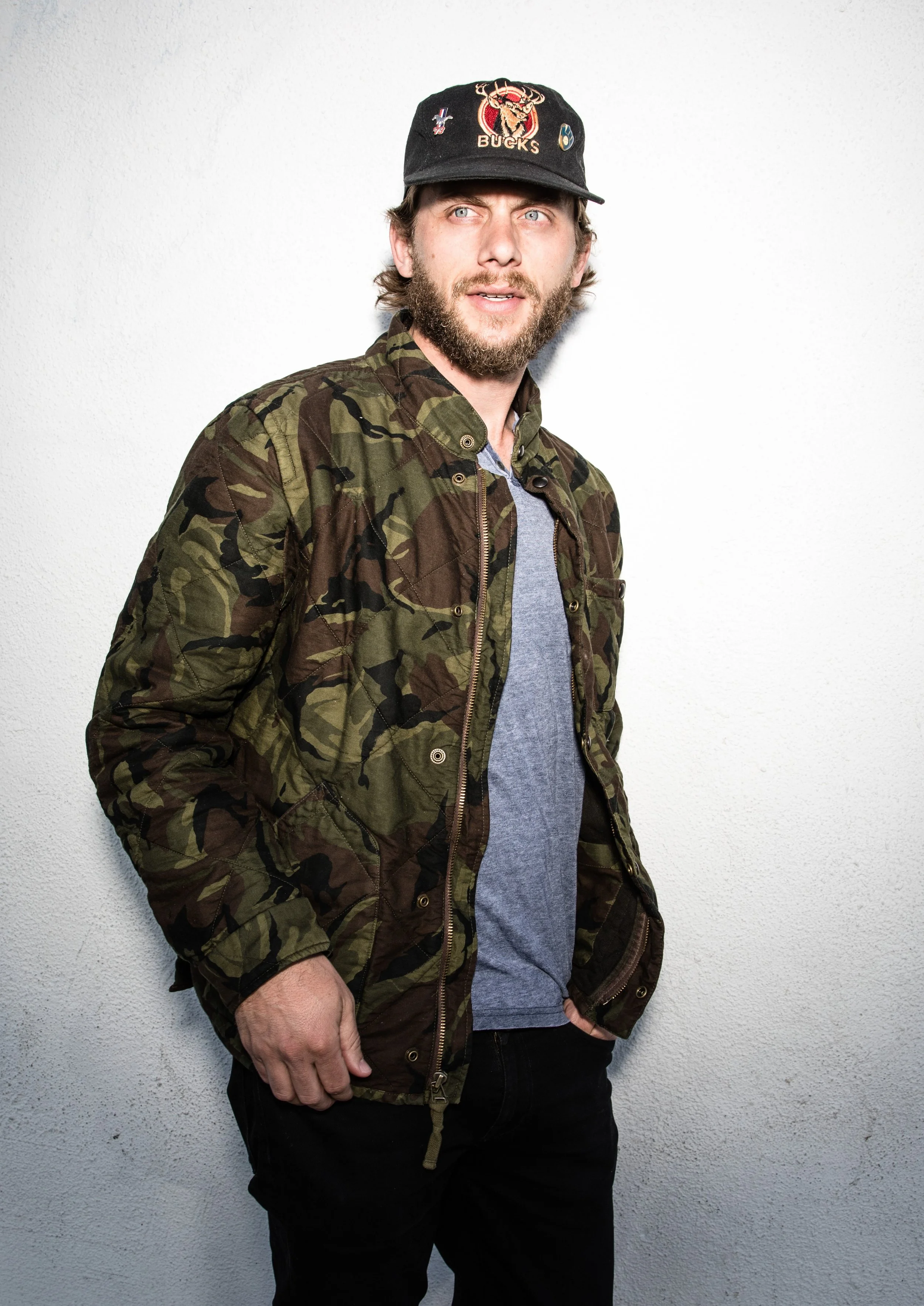 A man with light skin and a beard wearing a black Bucks cap, a camouflage jacket, and a gray shirt, standing against a plain white wall.