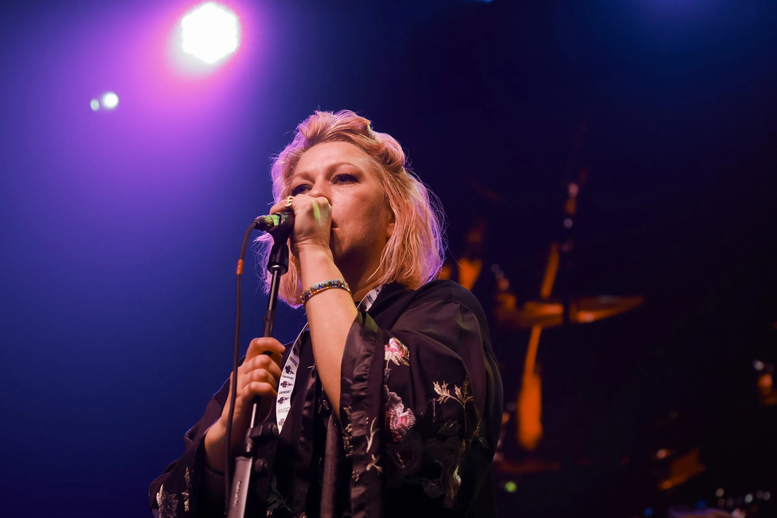 A woman singing into a microphone on stage, with colored stage lights illuminating her face.