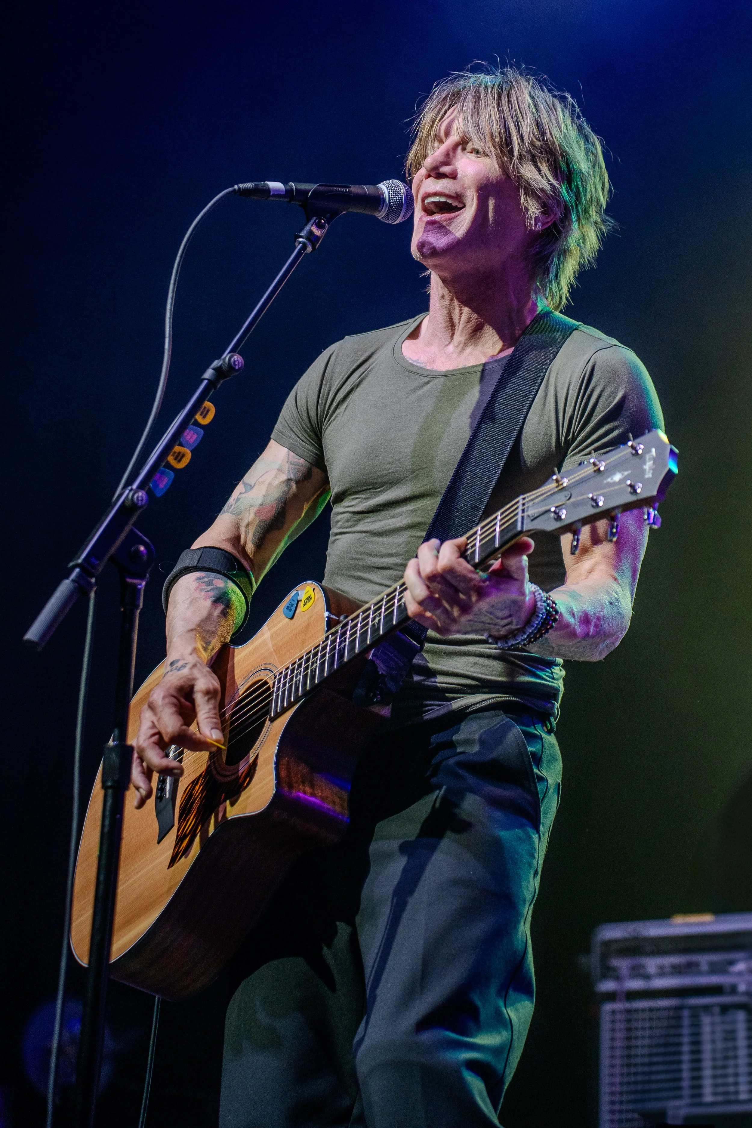 A man with long hair and tattoos plays an acoustic guitar and sings into a microphone on stage, with stage lights illuminating him.