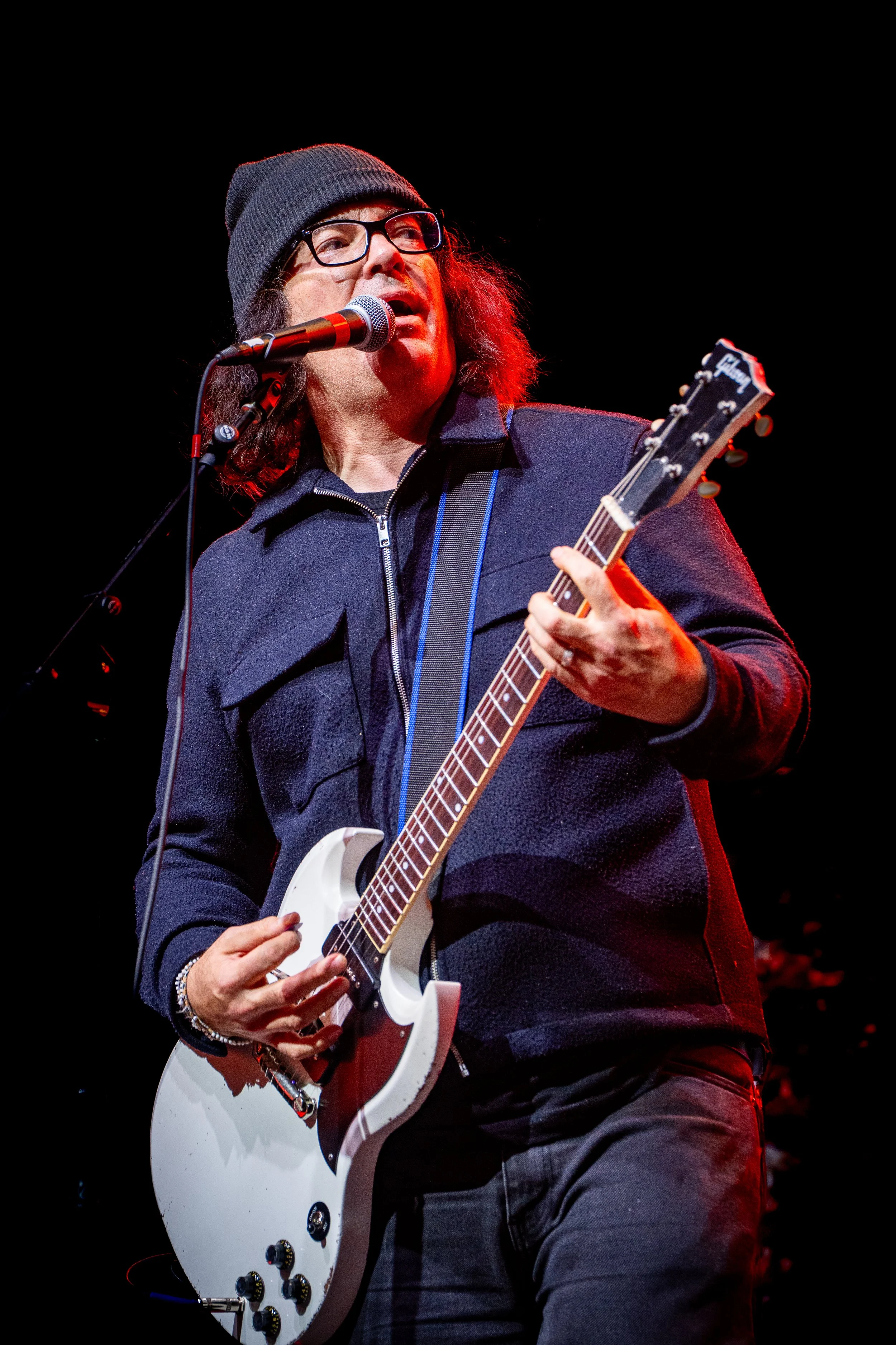 A man with long hair, glasses, a black beanie, and a dark jacket playing a white electric guitar and singing into a microphone on stage against a black background.