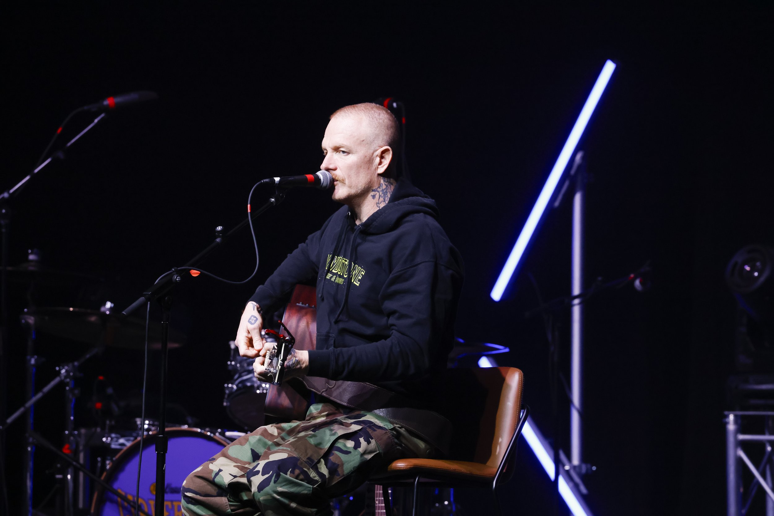 A man with a shaved head, tattoos on his neck and arms, wearing a black hoodie and camouflage pants, sitting on a chair and playing an acoustic guitar on a dark stage with blue neon lights.