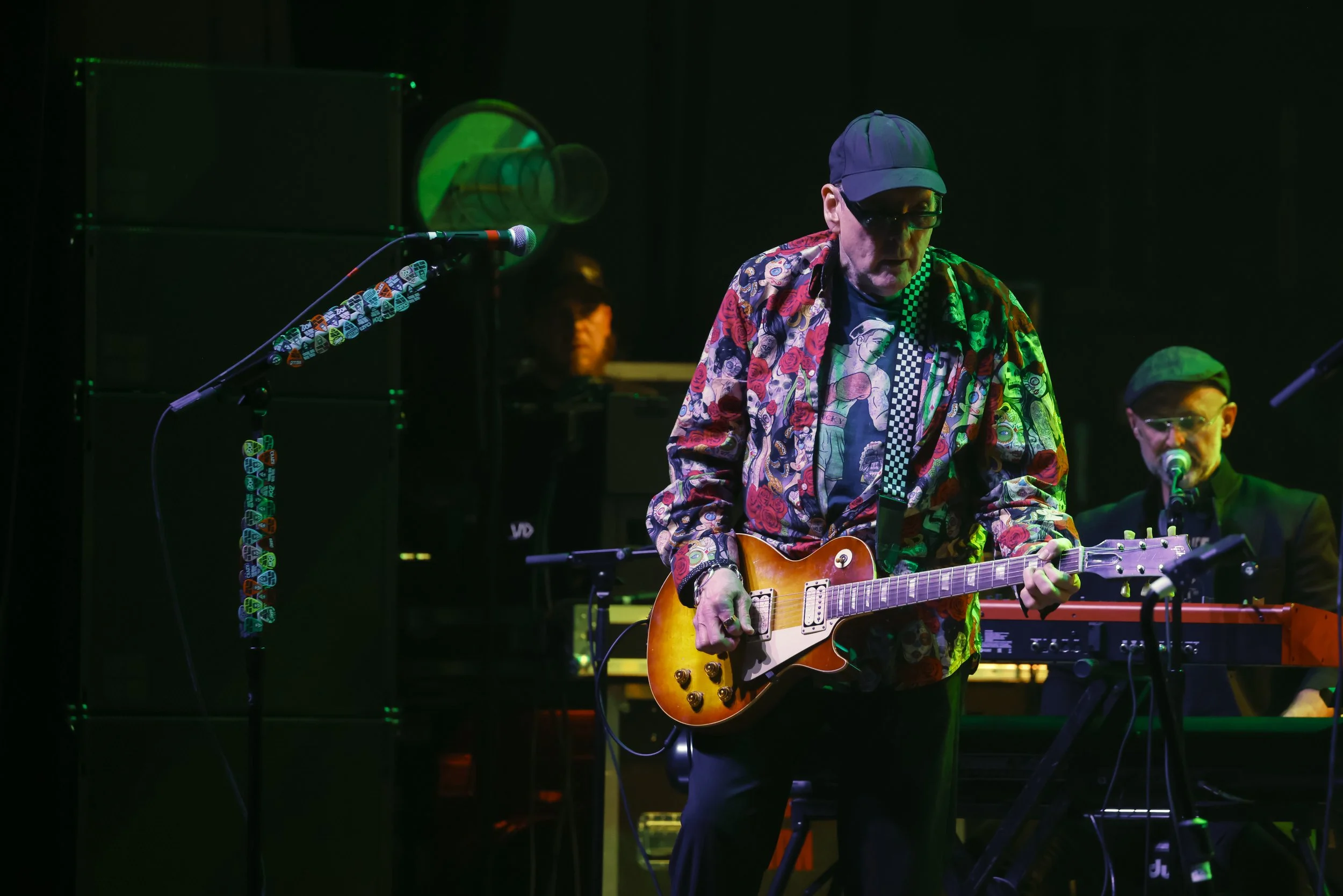 Musician playing an electric guitar on stage with a keyboard and microphone in the background, wearing a colorful patterned shirt and cap, with stage lighting.