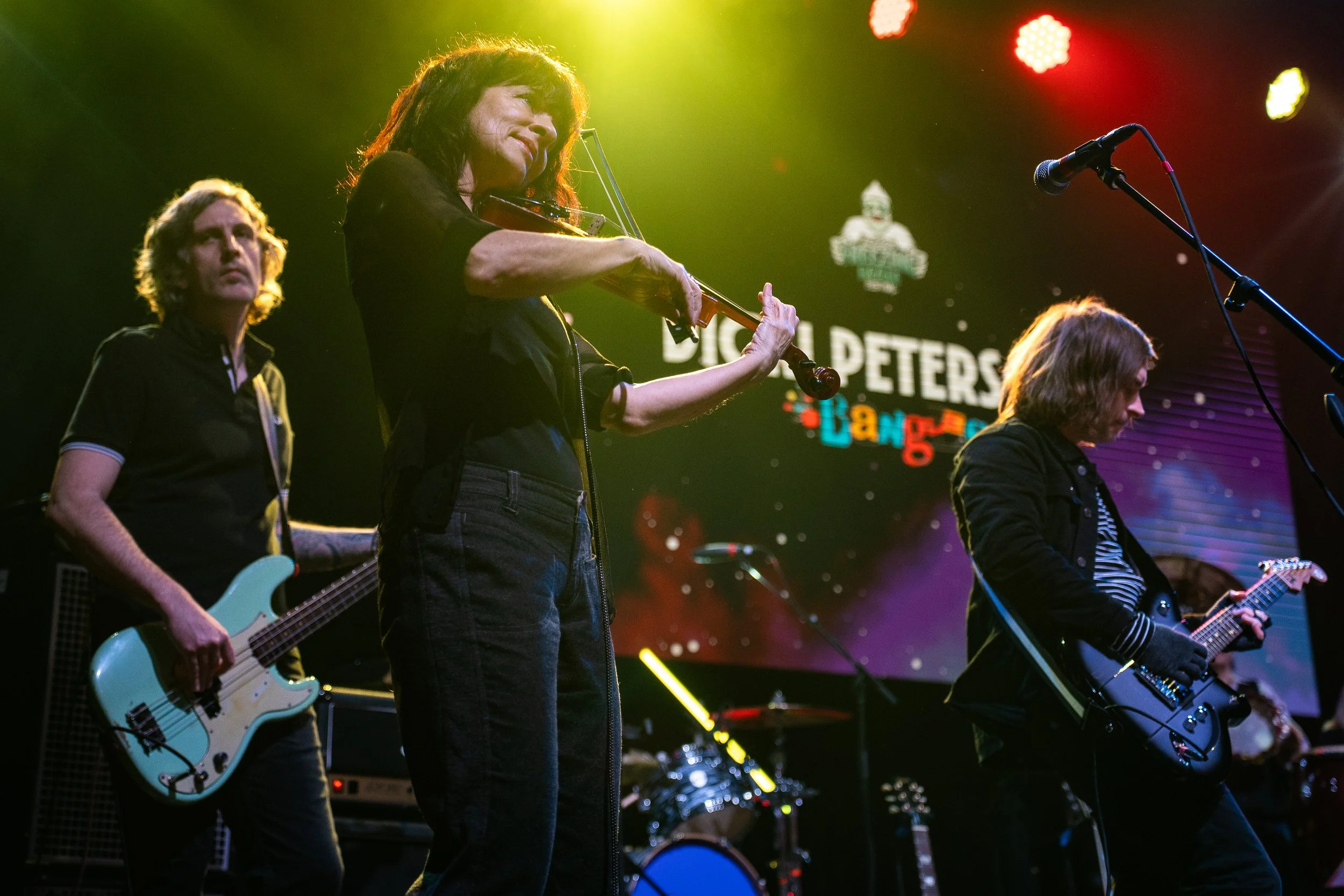 Four musicians performing on stage: a bassist, a violinist, and two guitarists, under colorful stage lights, with a digital backdrop.