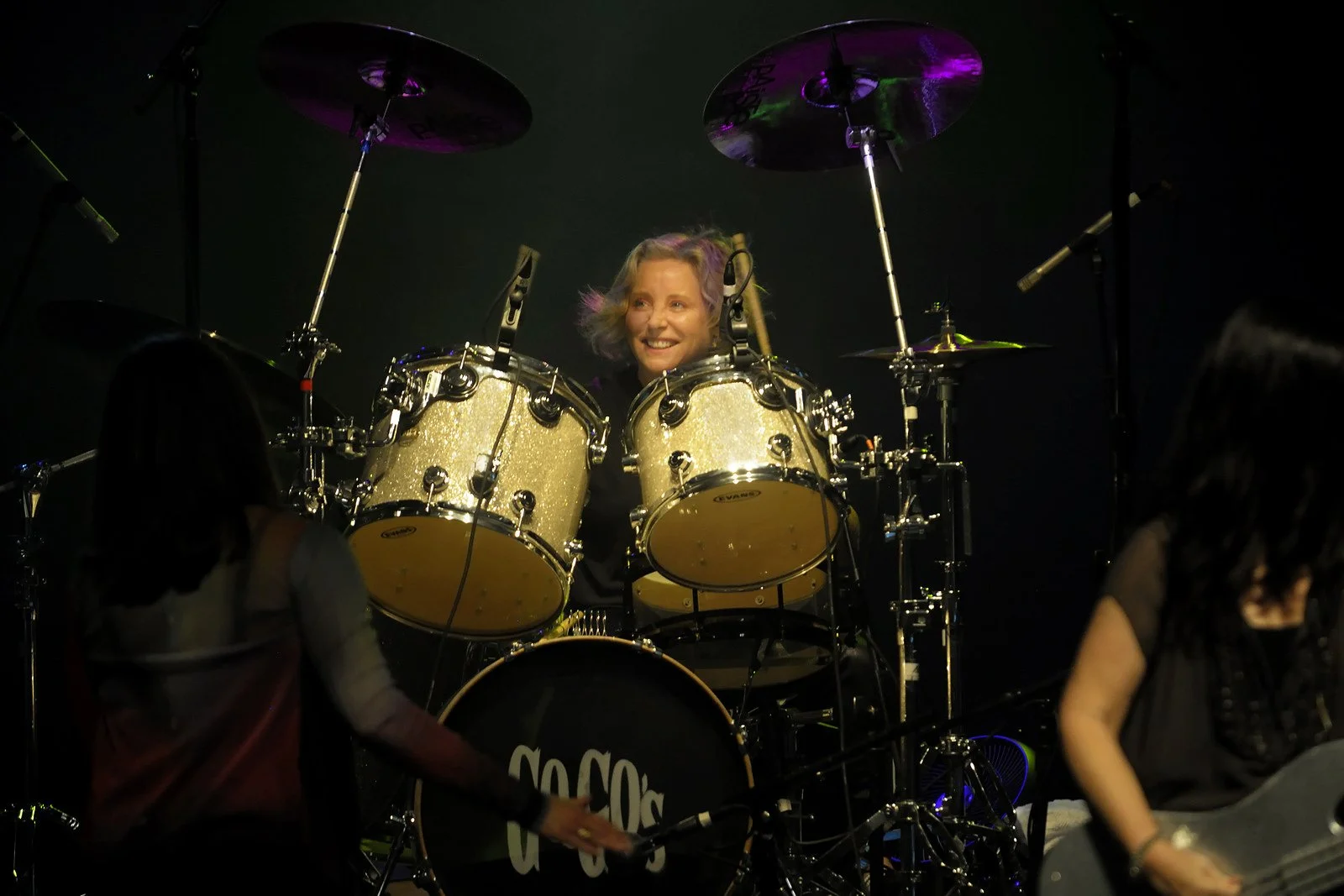 A woman with curly blonde hair smiling while sitting behind a gold drum set on stage, with music stands and other performers around, in a dark venue with stage lighting.