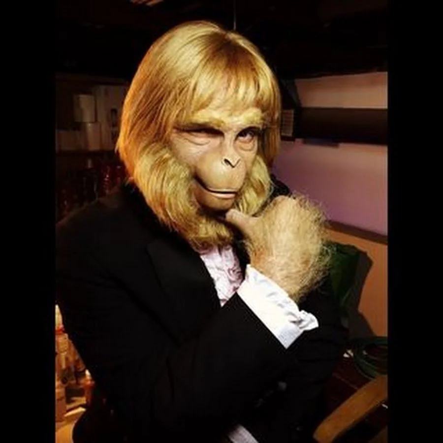 Person wearing a monkey mask with blonde hair and furry hand, dressed in a black jacket and white shirt, posing with one hand on chin in a room with soft lighting.