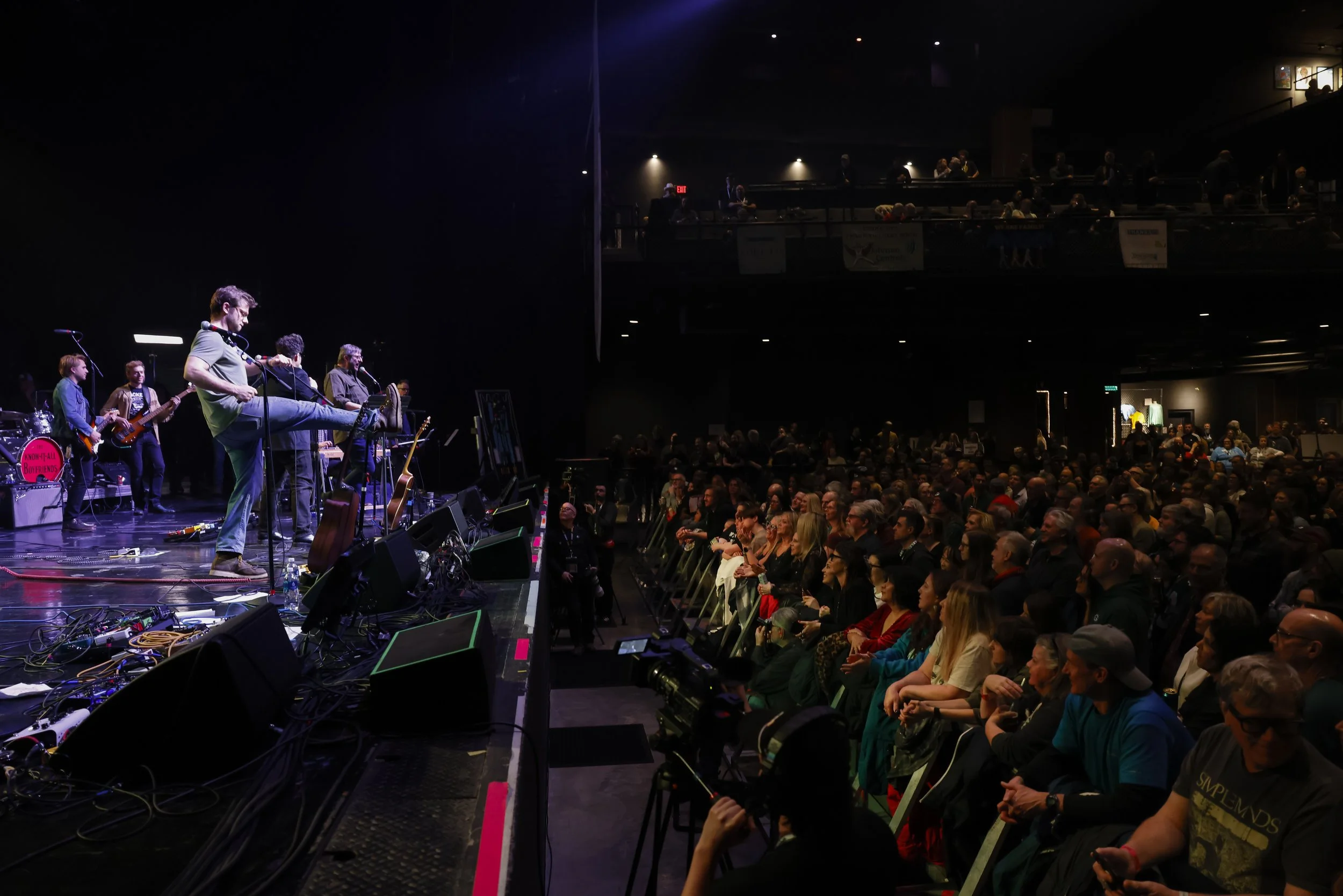 Concert stage with musicians performing and an audience watching in a dark theater, illuminated by stage lights.