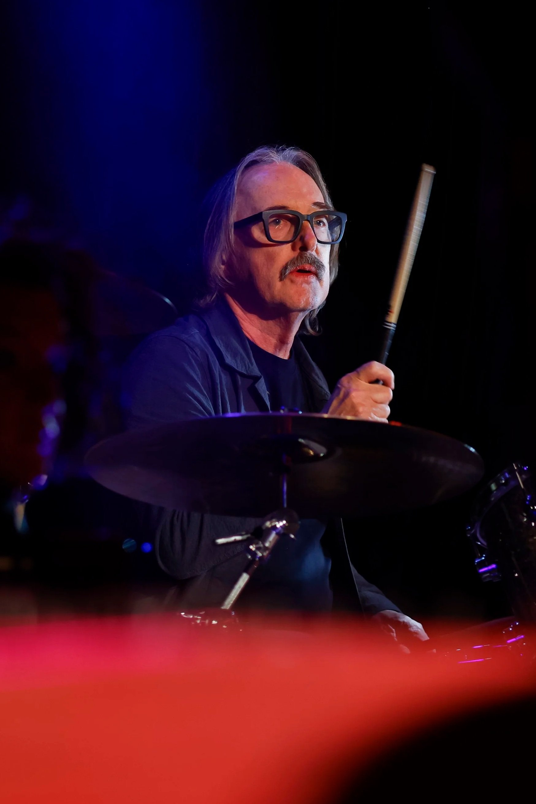 A man with gray hair, thick glasses, and a mustache, wearing a black shirt, sitting in front of a dark background.