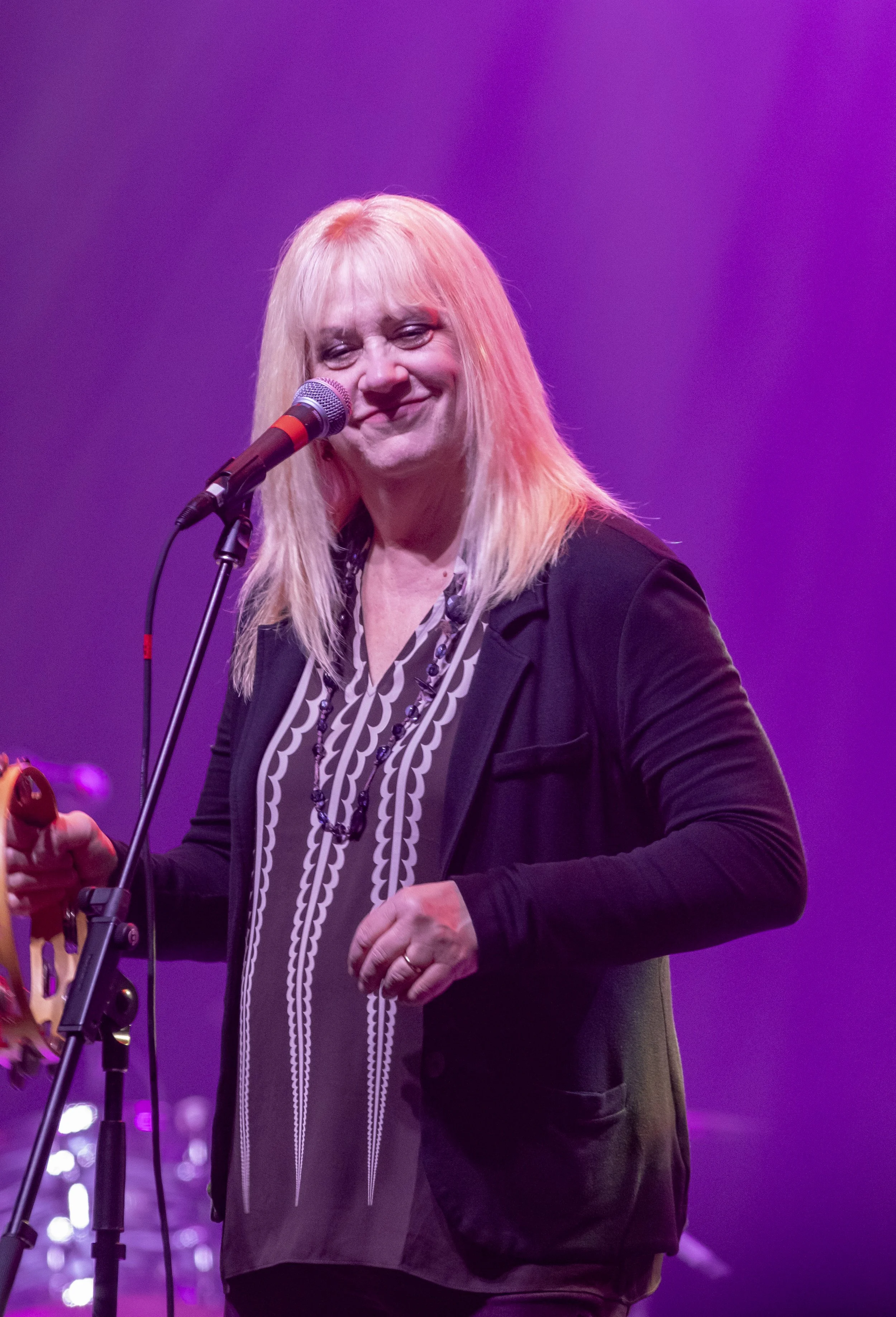 A woman with long blonde hair smiling while singing into a microphone on stage with purple lighting.