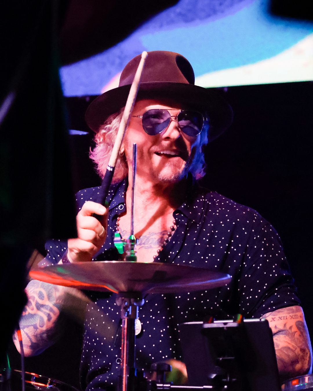A drummer with long hair wearing sunglasses and a wide-brimmed hat, smiling while playing the drums with a drumstick in hand.