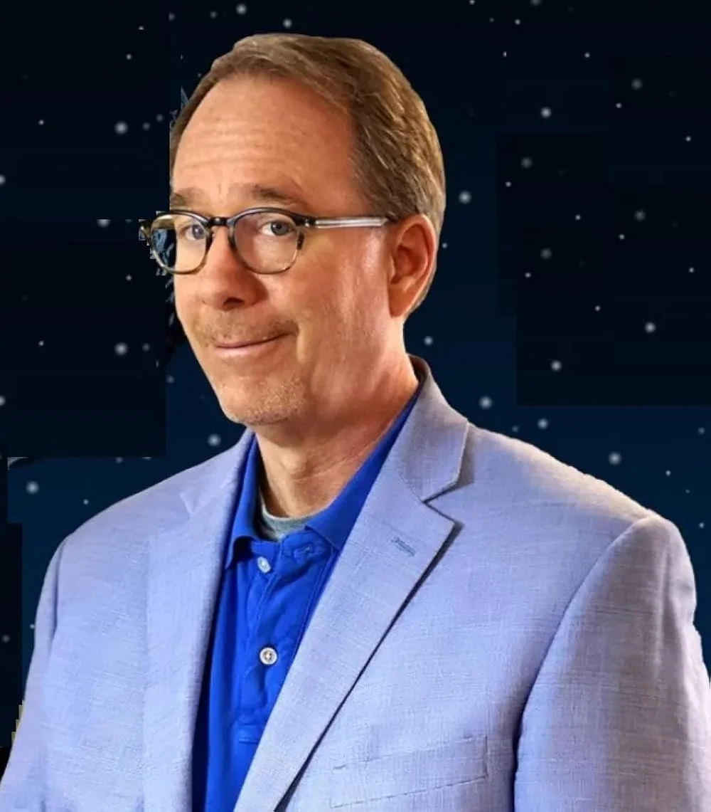 A man with glasses, wearing a light gray blazer and blue shirt, standing against a starry night sky background.