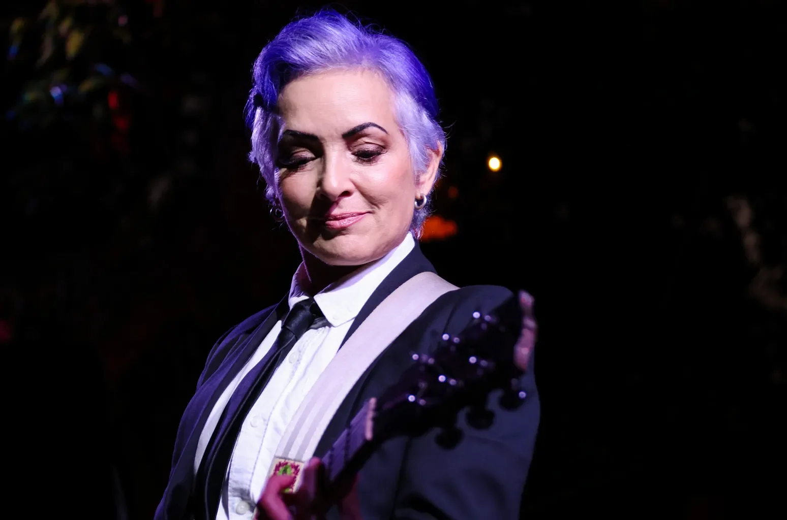 An elderly woman with short gray hair, wearing a black blazer with decorative buttons on the shoulder, a white shirt, and a black tie, playing an electric guitar with a passion.
