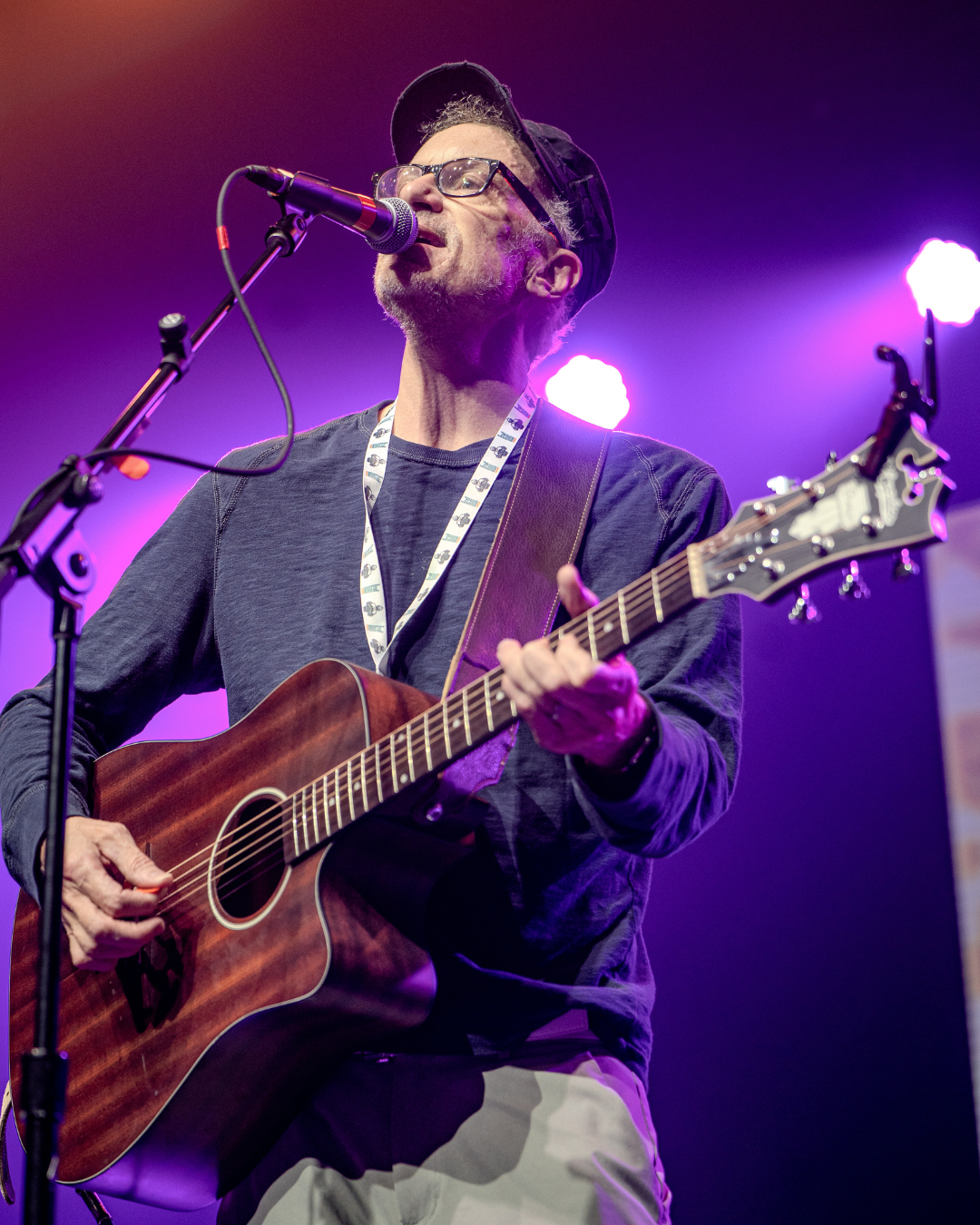 A man wearing glasses and a baseball cap is playing an acoustic guitar and singing into a microphone on stage, with colorful stage lights in the background.