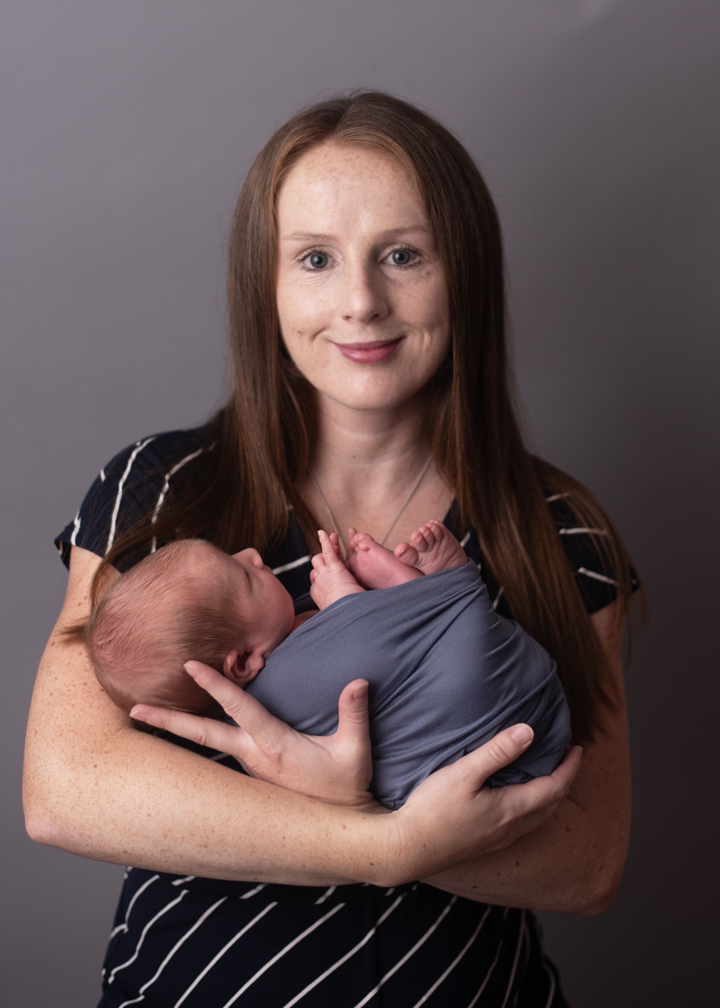 A woman with red hair and freckles holding a sleeping newborn baby wrapped in a gray blanket, against a gray background.