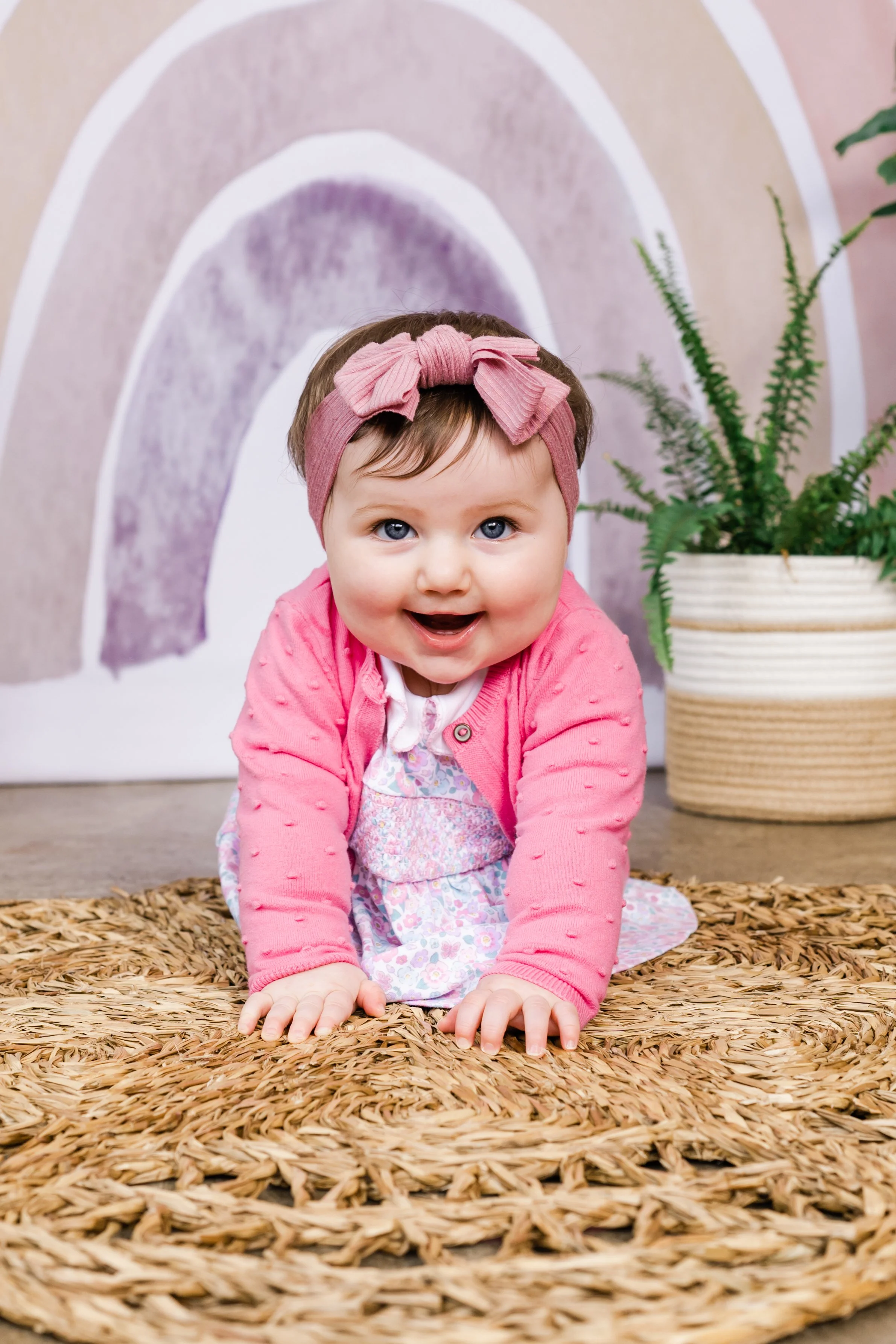 A smiling baby girl is crawling on a textured brown rug with a pink headband and a pink cardigan. Behind her, there is a decorative rainbow wall art and a large potted green fern plant.
