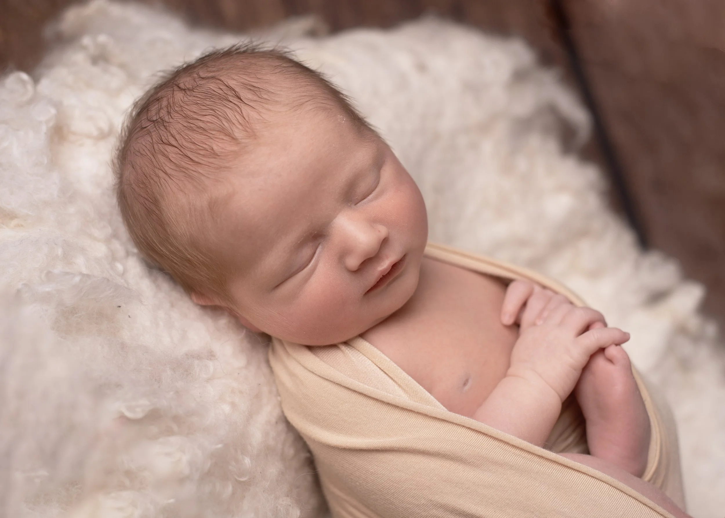 Close-up of a sleeping newborn baby lying on a fluffy white blanket, wearing a light peach-colored outfit.