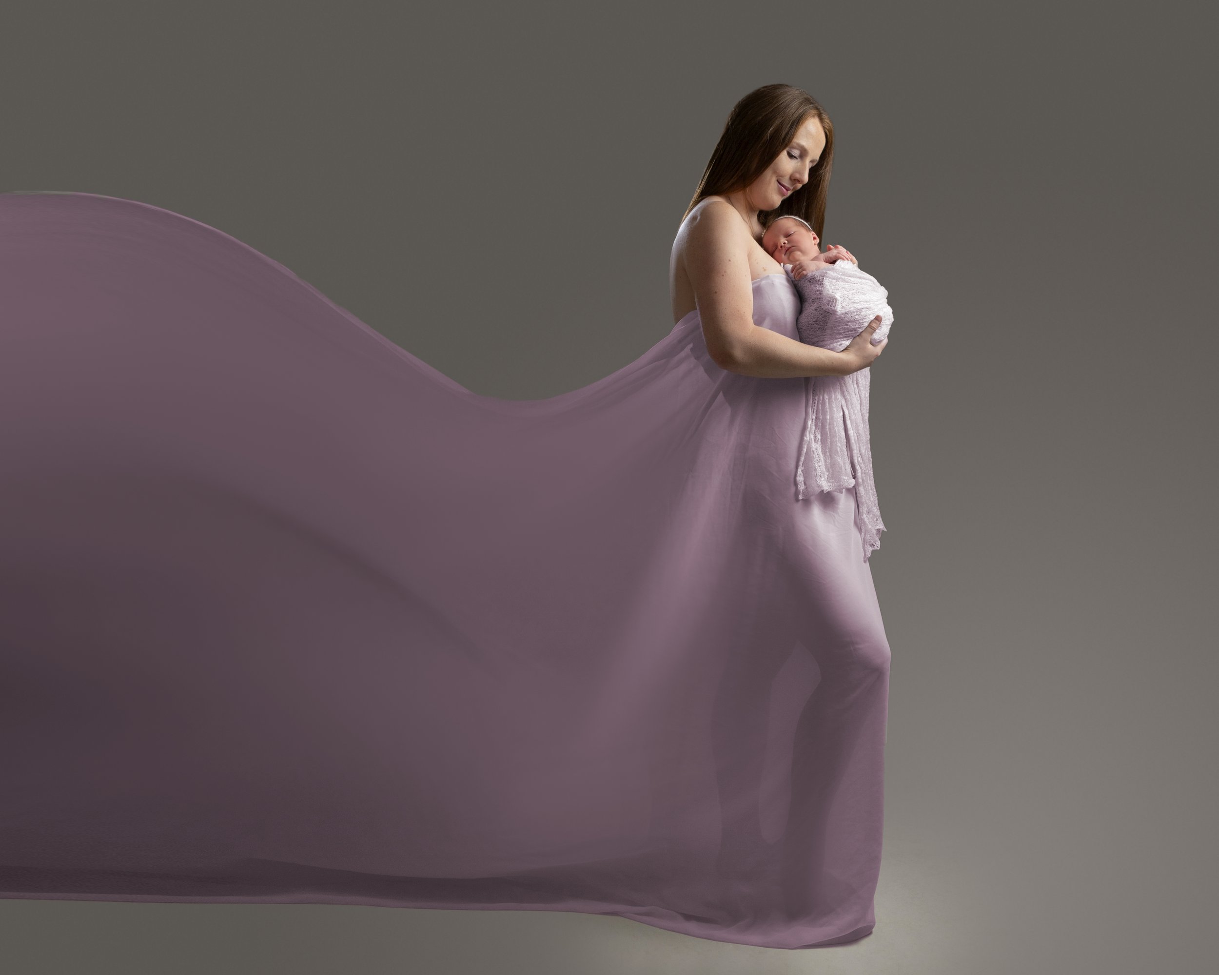 A woman with long brown hair, wearing a strapless pink dress, holds a newborn baby wrapped in a gray blanket, against a plain gray background.