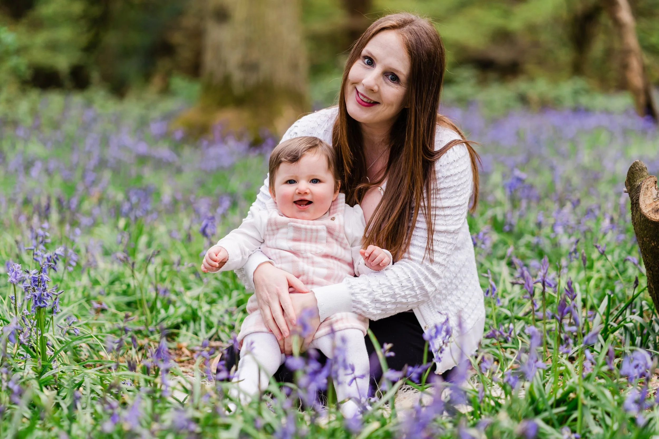 A woman with long red hair and a baby girl sitting on the ground in a field of purple flowers, smiling at the camera.