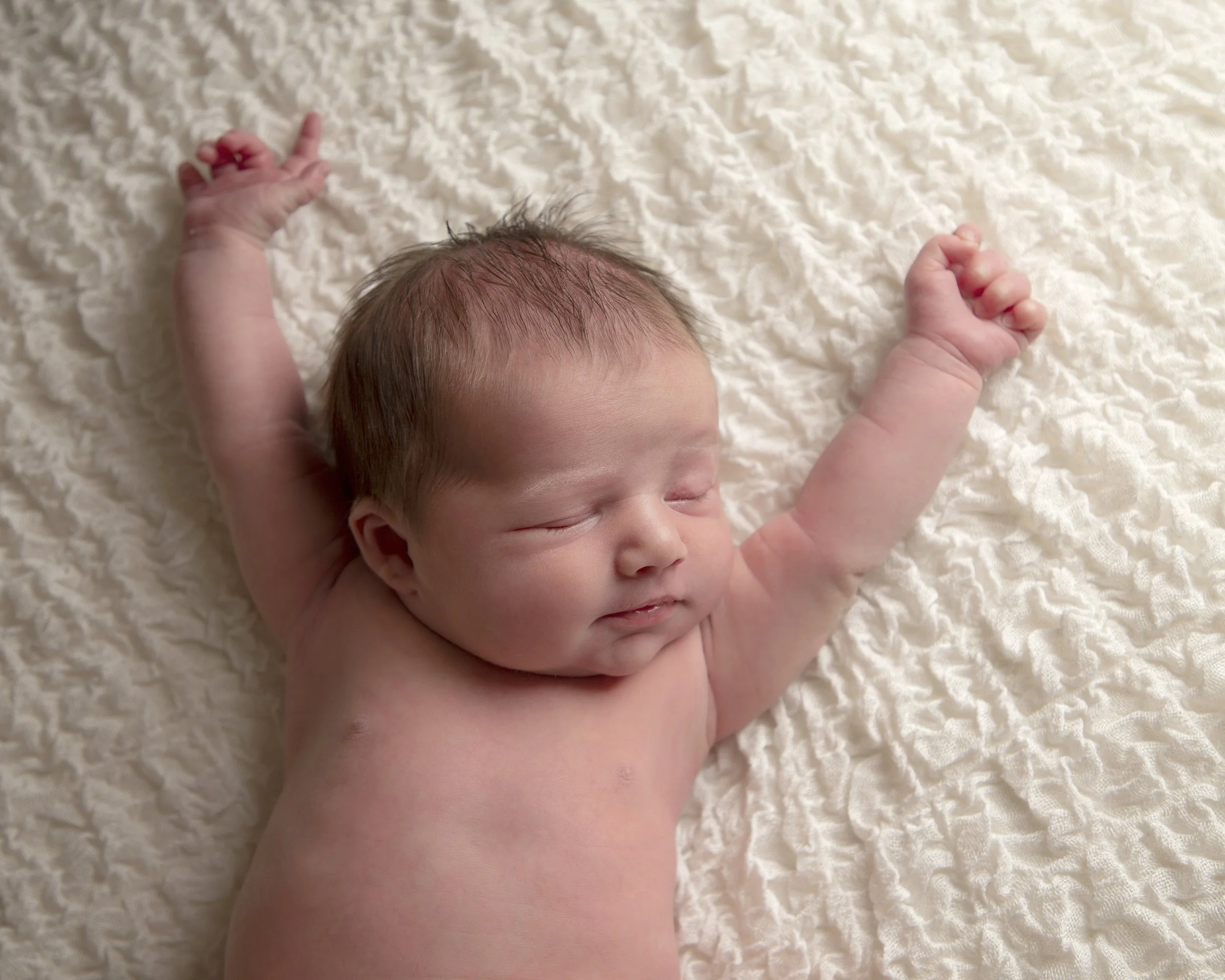 Close-up of a sleeping baby with light skin and brown hair, lying on a soft, textured, cream-colored blanket, with arms raised.