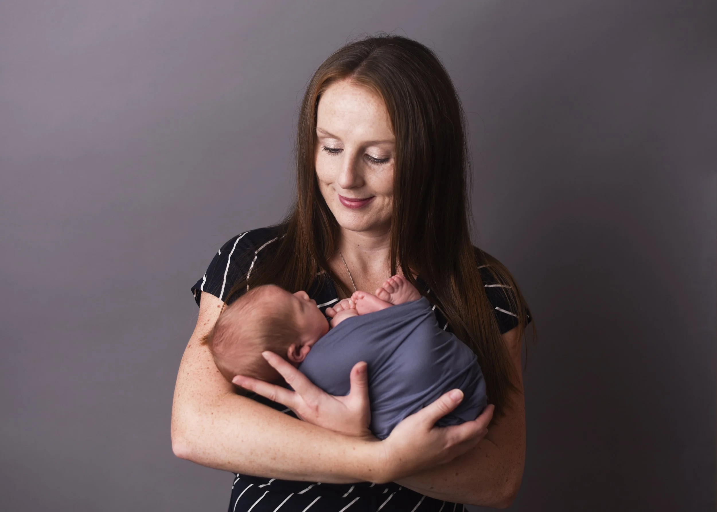 A woman with long brown hair and freckles is holding a baby, who appears to be a few months old, in her arms. The woman is looking down at the baby with a gentle smile. The background is plain gray.