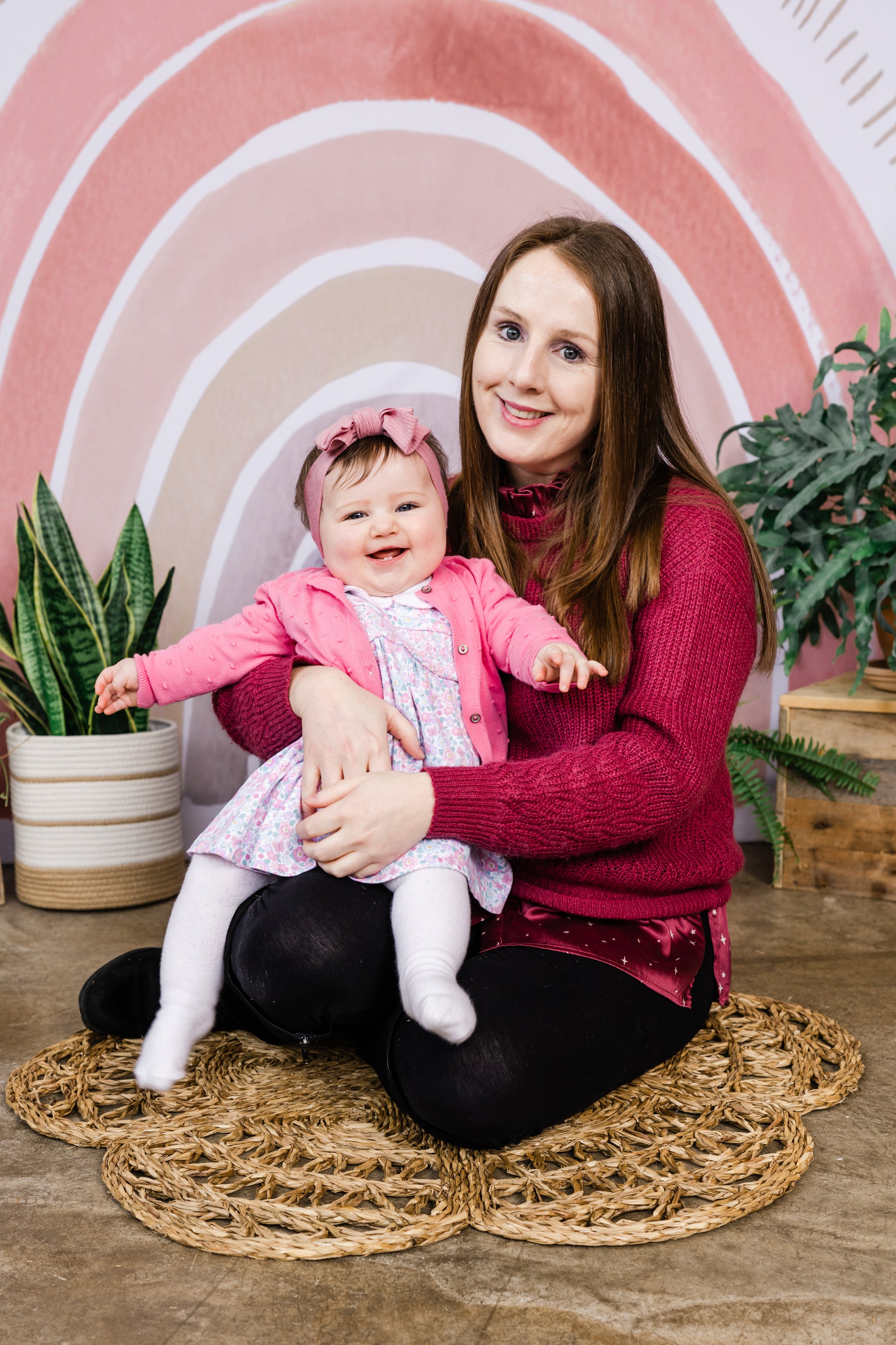 A woman with long brown hair and a young girl with a pink headband and pink sweater sitting on a woven rug with greenery and a rainbow backdrop.
