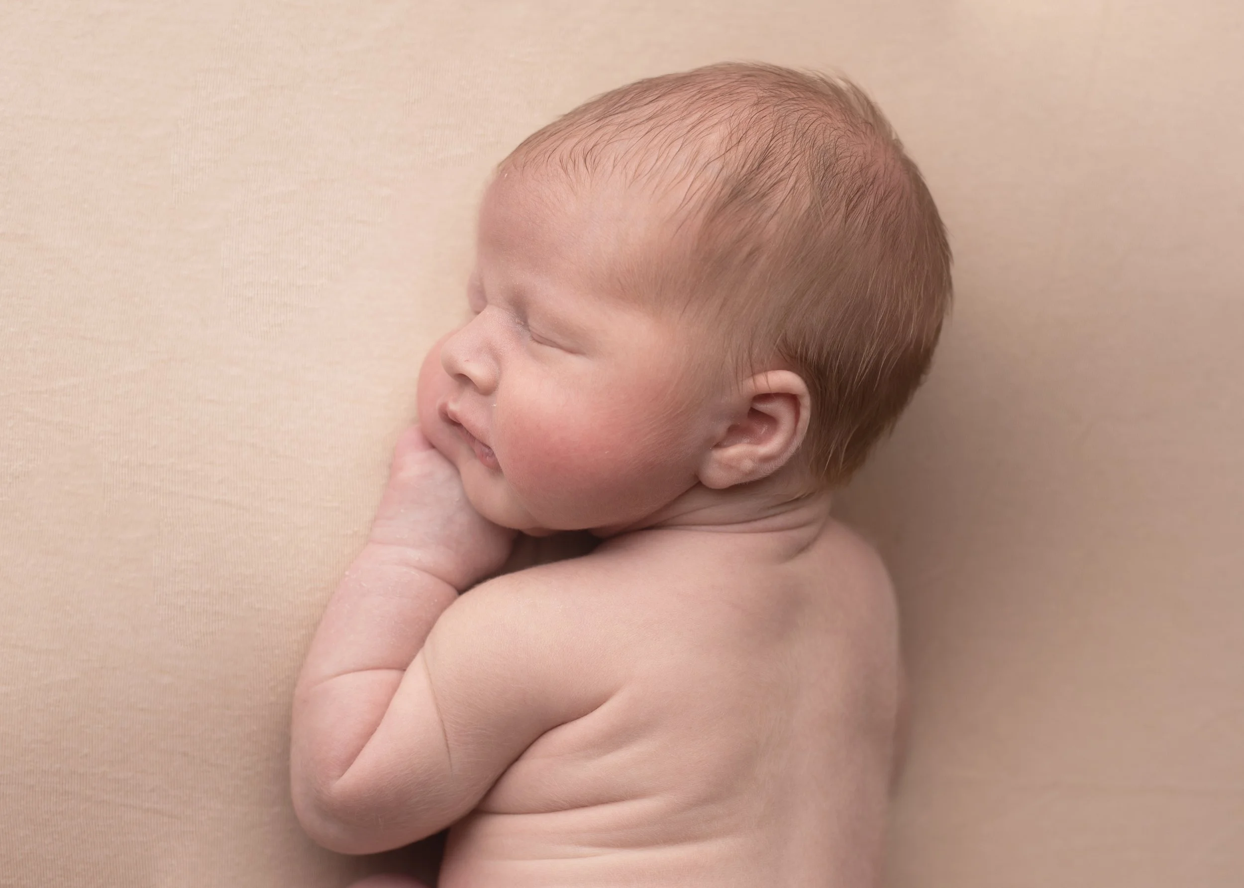 Close-up of a sleeping baby with light skin and short, reddish hair, lying on a soft, beige surface.