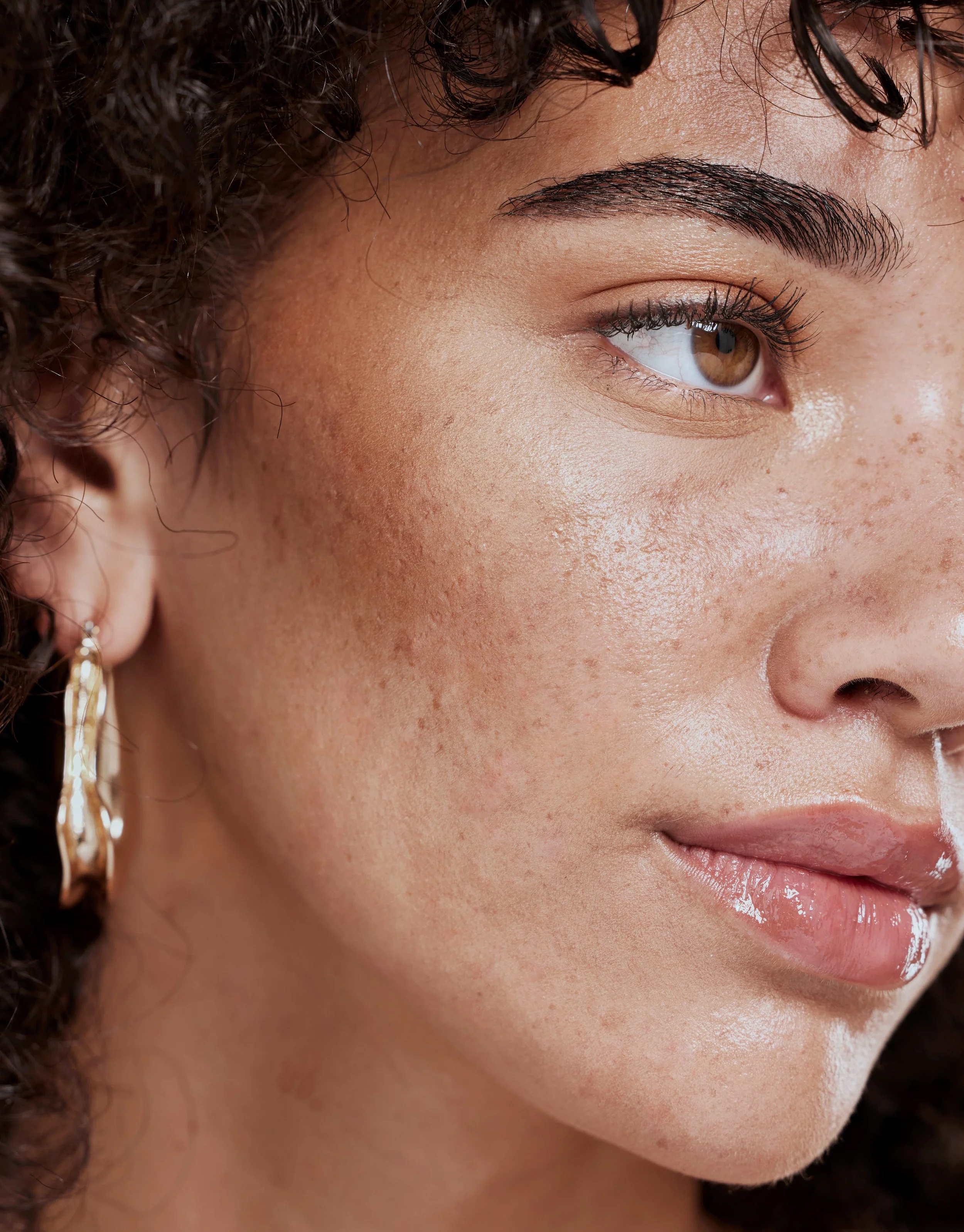 Close-up of a woman's face showing her eye, eyebrow, skin, and part of her lips and ear with a gold hoop earring. Her brown eye is partially open, with long eyelashes and well-defined eyebrows. Her skin is smooth with freckles visible.