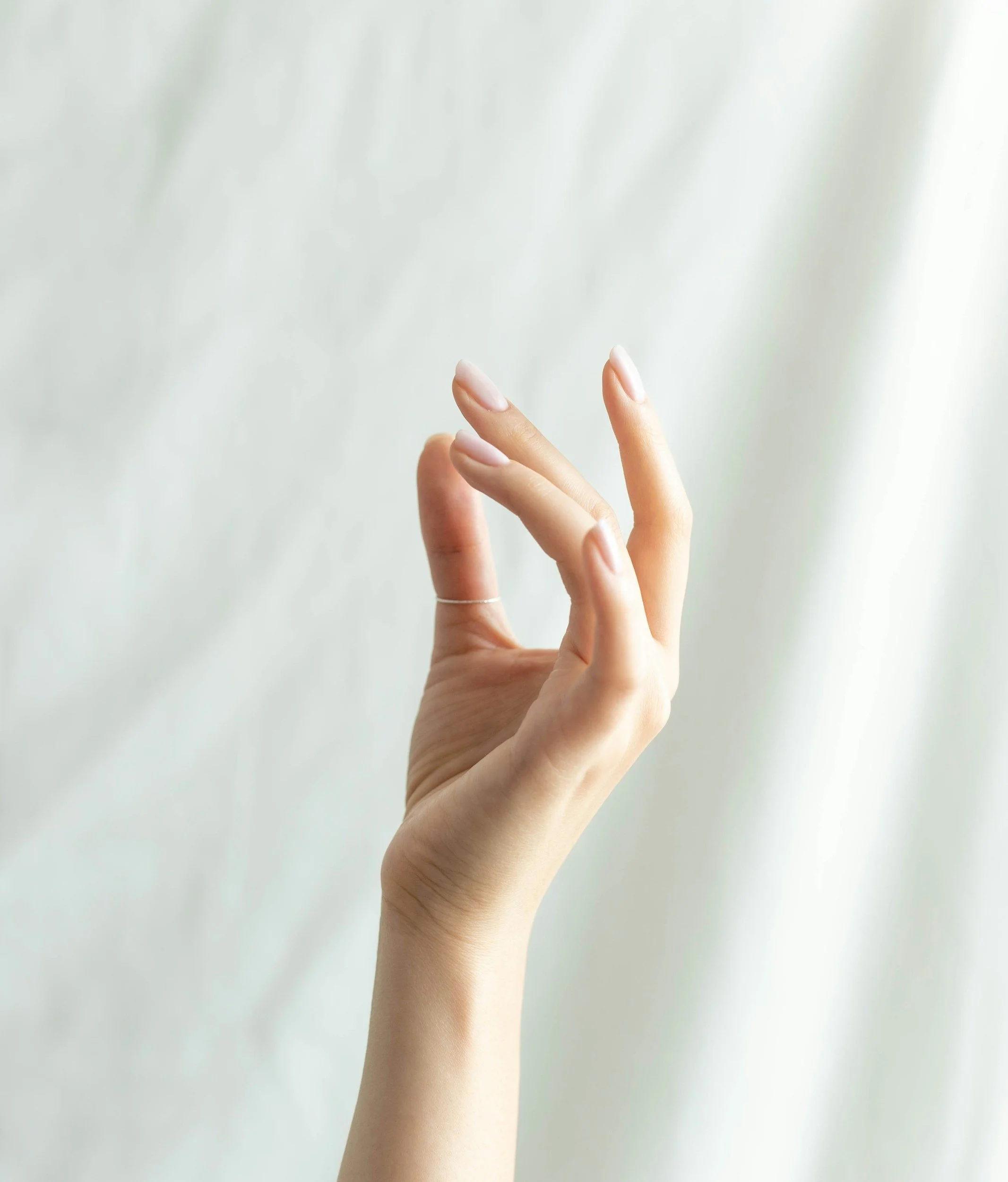 A person's hand with well-manicured nails and a thin ring, gently touching a semi-transparent white curtain.