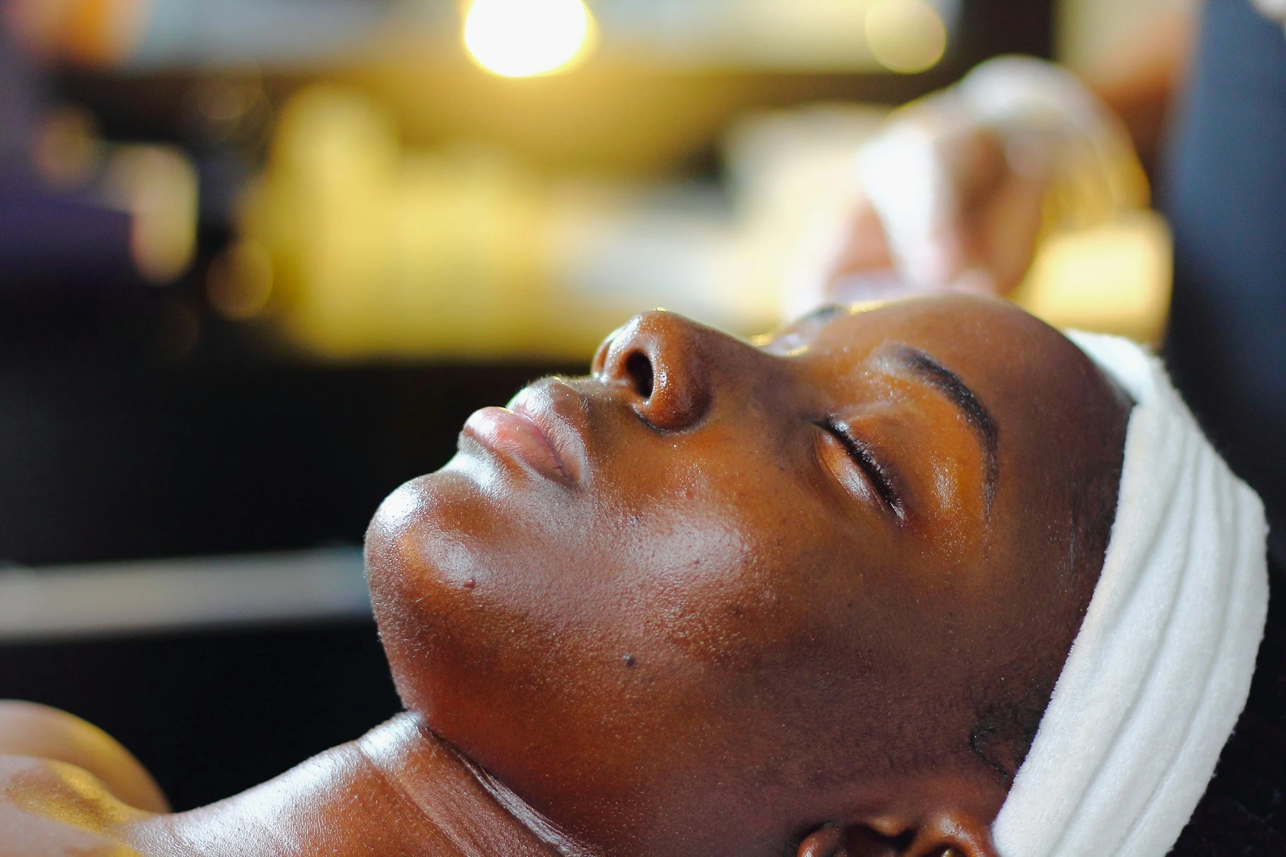 Close-up of a woman with closed eyes, her face turned upward, possibly receiving a massage or facial treatment, with warm, blurred lights in the background.