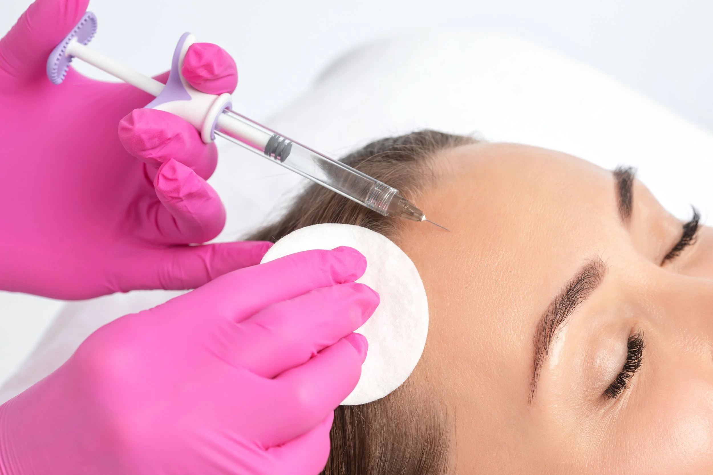 A person receiving a cosmetic injection in their forehead, with the practitioner wearing pink gloves and holding a syringe. The person's face is relaxed, lying on a white pillow.