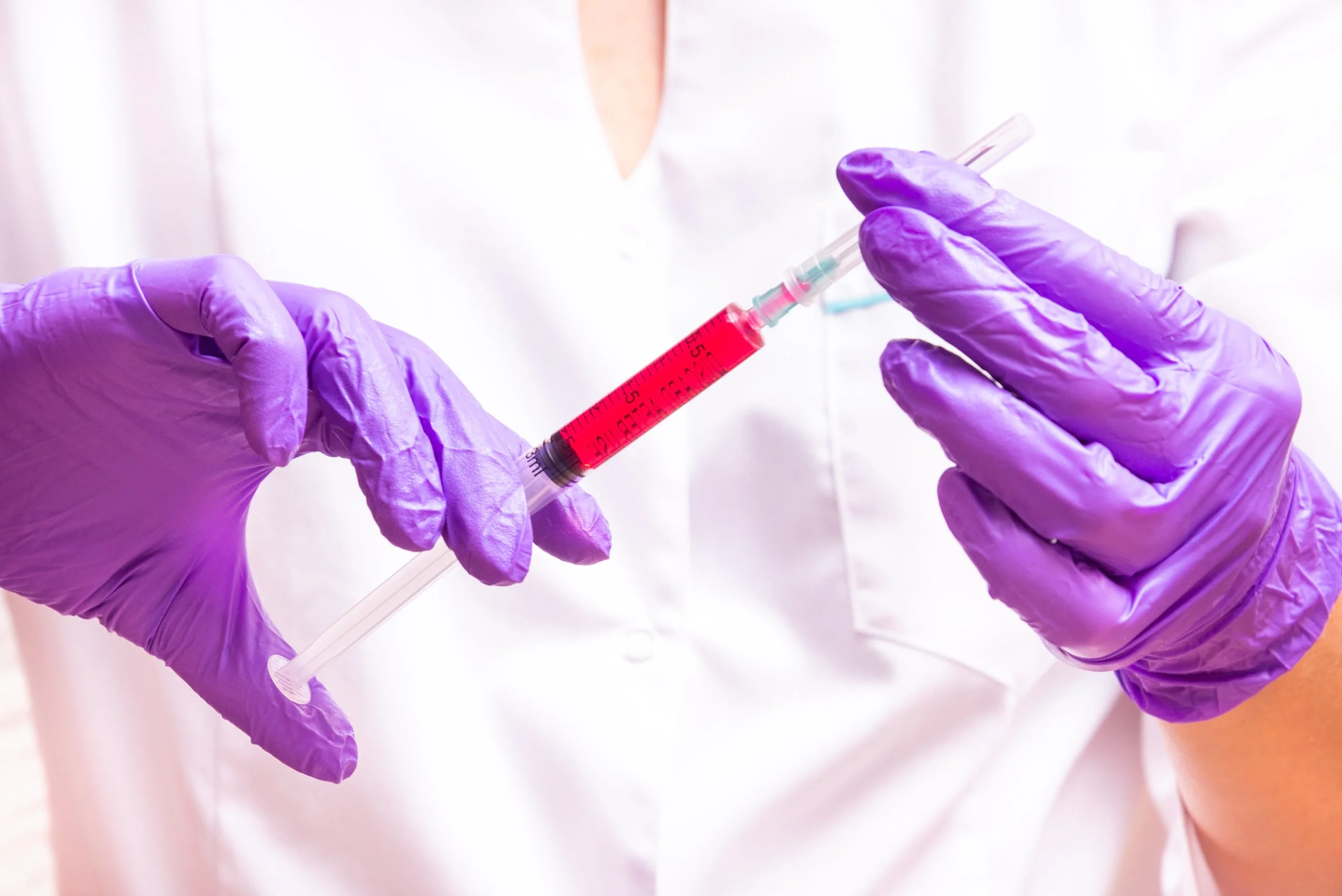 A healthcare professional wearing purple gloves drawing red liquid into a syringe.