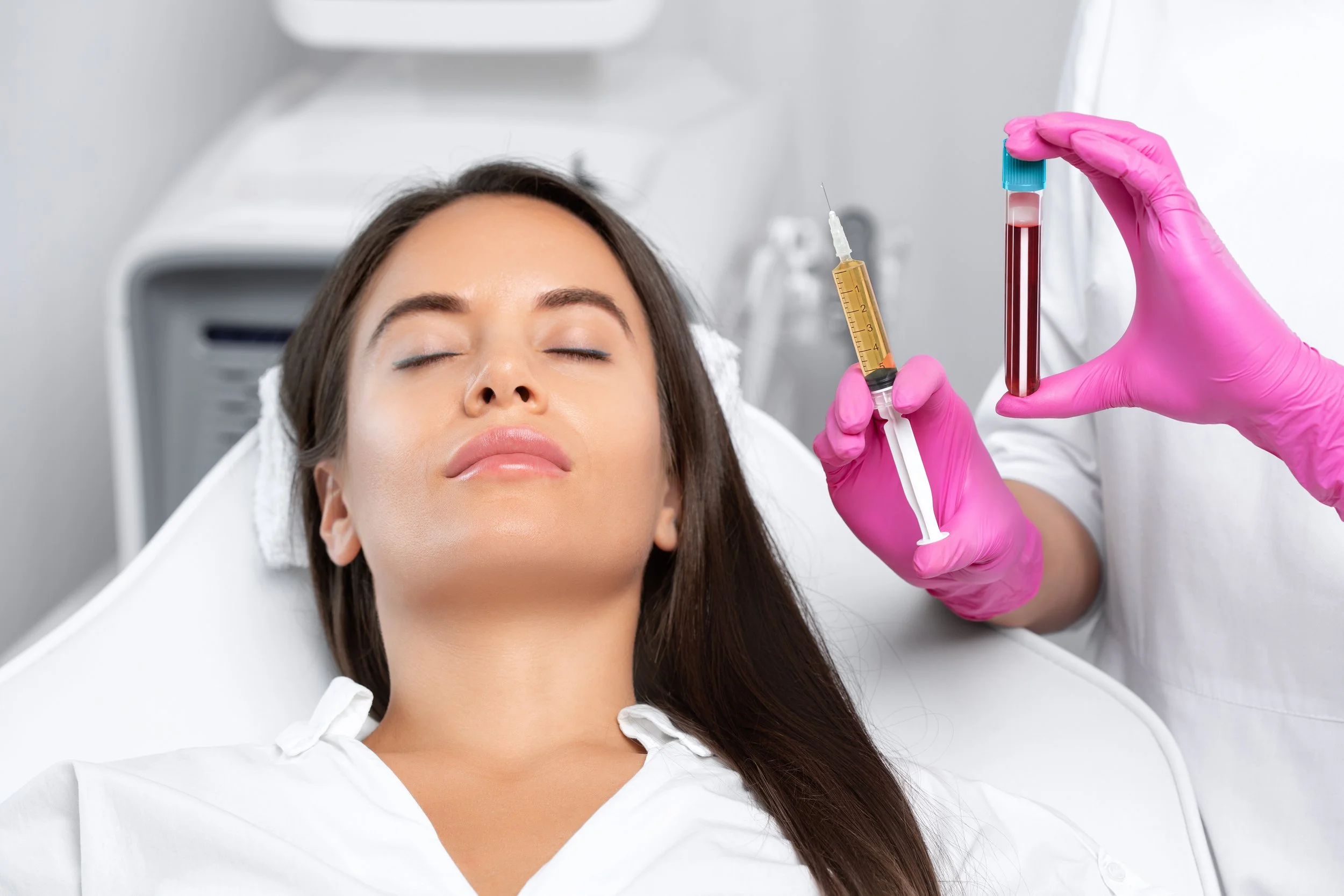 A woman is lying down with eyes closed, receiving a blood draw from a healthcare professional wearing pink gloves. The healthcare professional is holding a test tube filled with blood and a syringe with a needle.