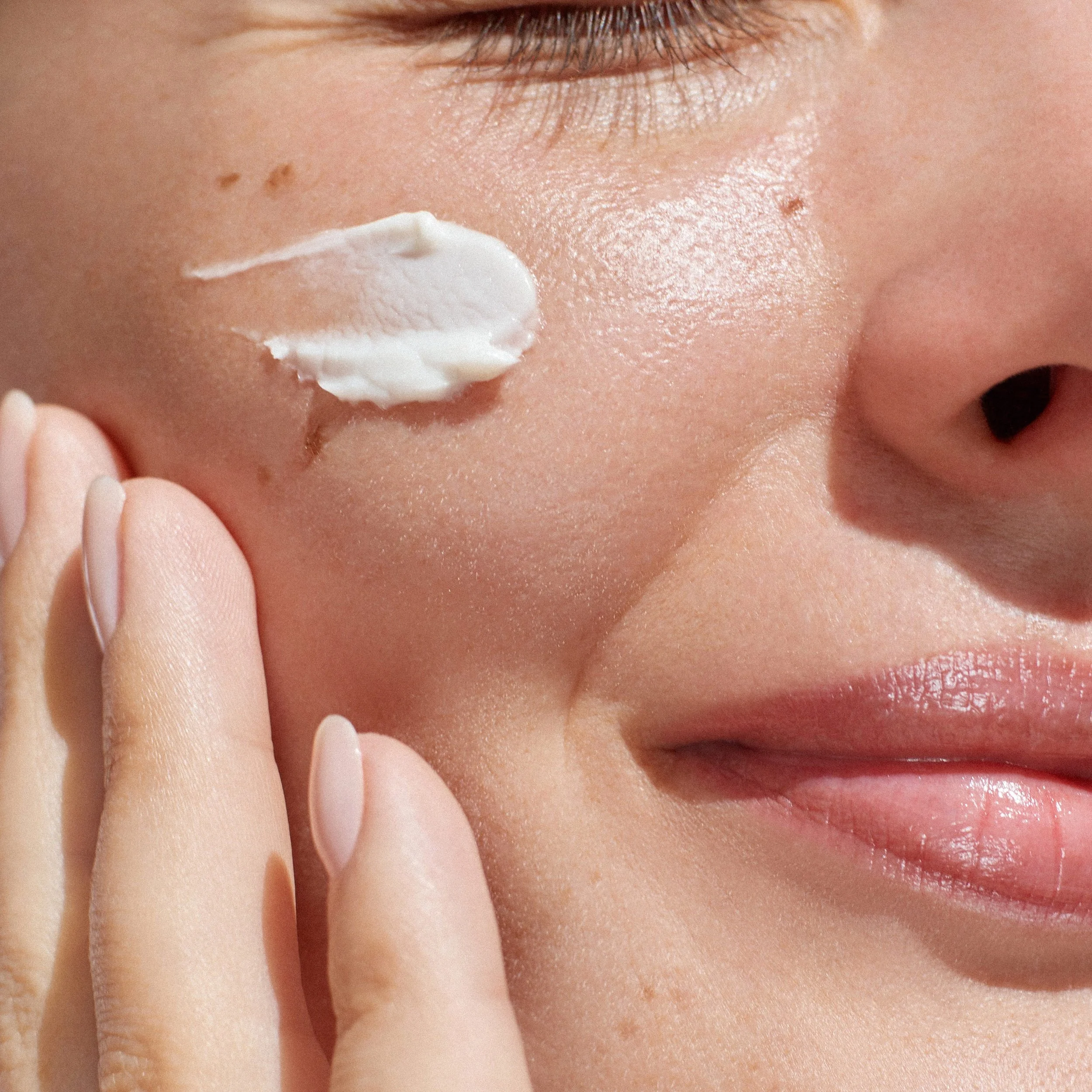 Close-up of a person's face with an eye closed, applying cream to the nose with fingers.