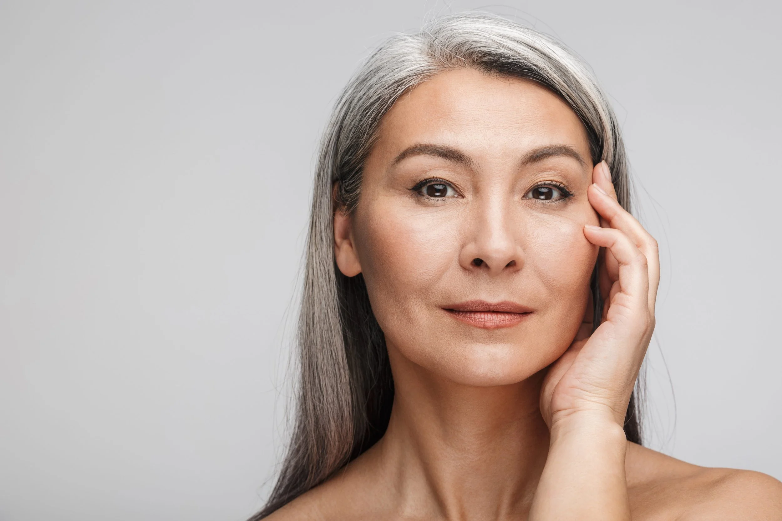 Close-up of a mature woman with long gray hair touching her face with her hand, against a plain light gray background.