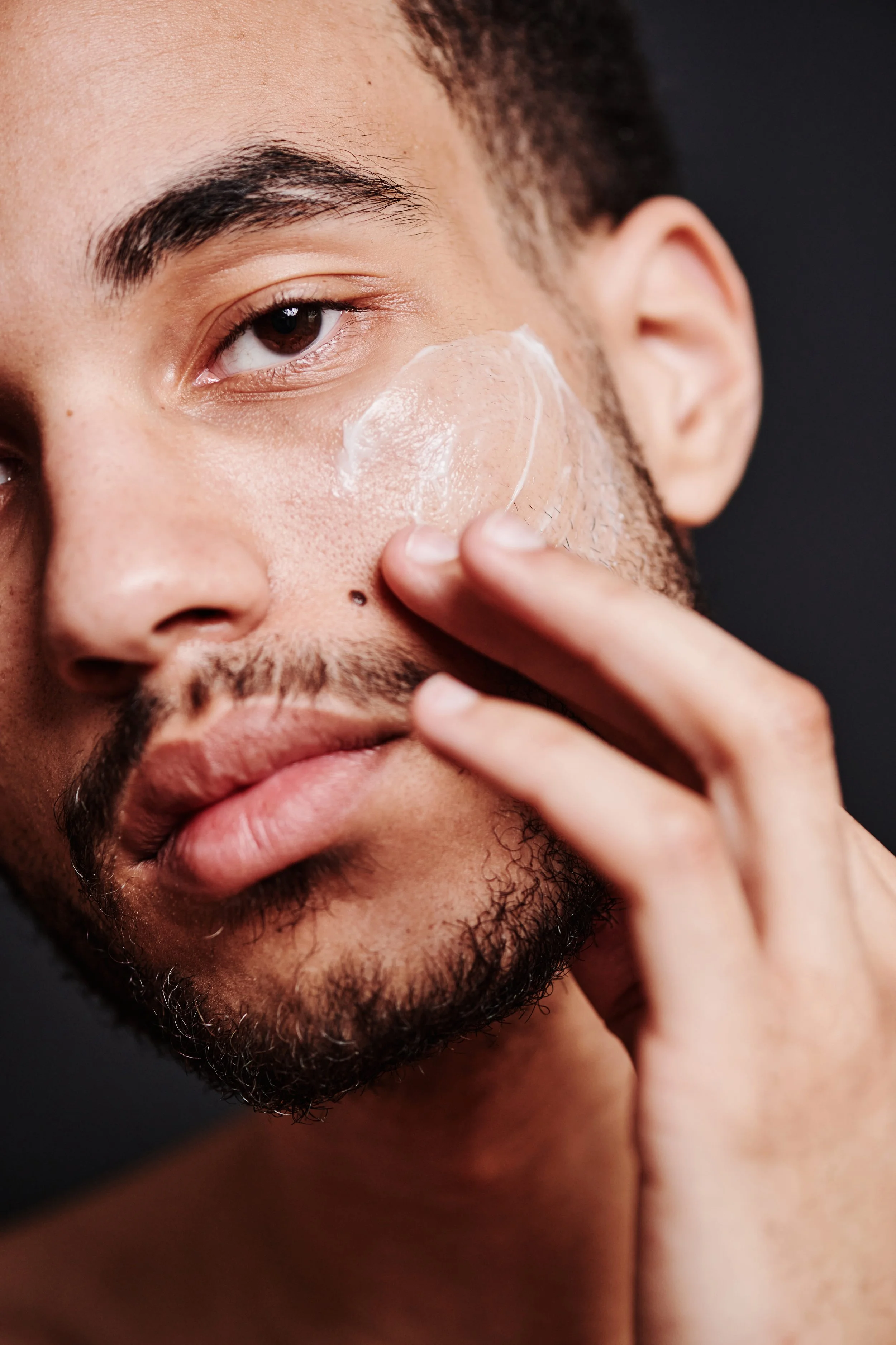 A close-up of a young man's face as he applies a clear facial mask or skincare product to his cheek with his finger.