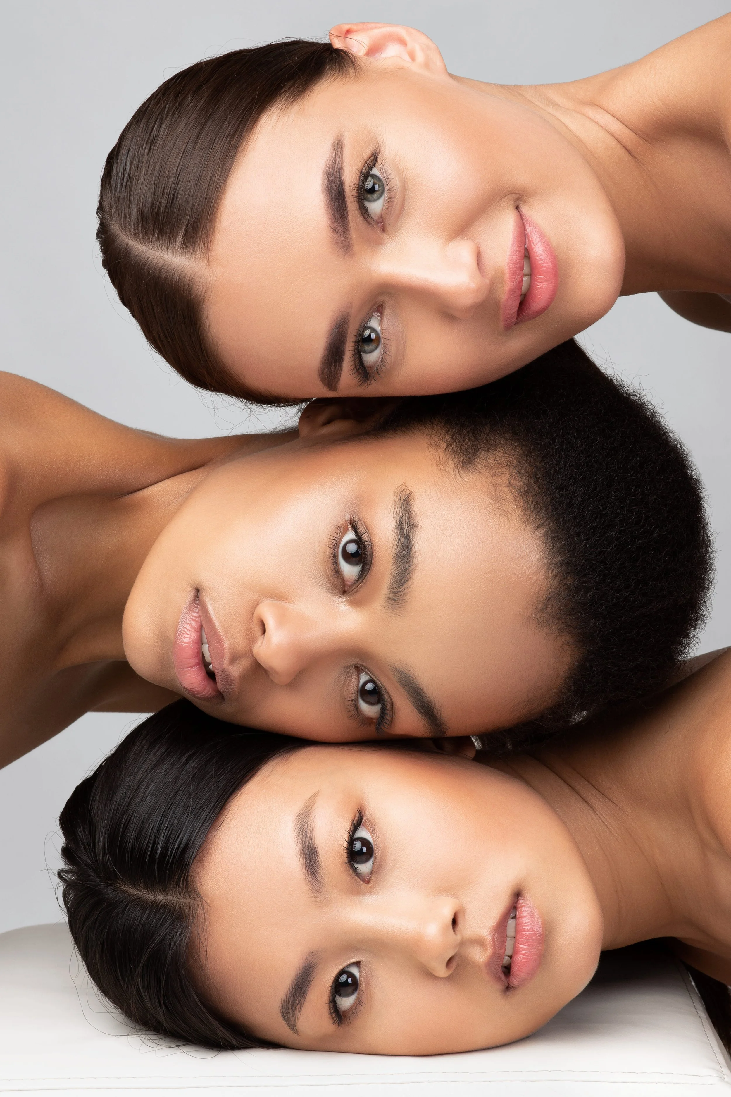 Four women with diverse skin tones and different hairstyles are closely positioned, lying down with their heads touching. They are looking directly at the camera with neutral expressions.