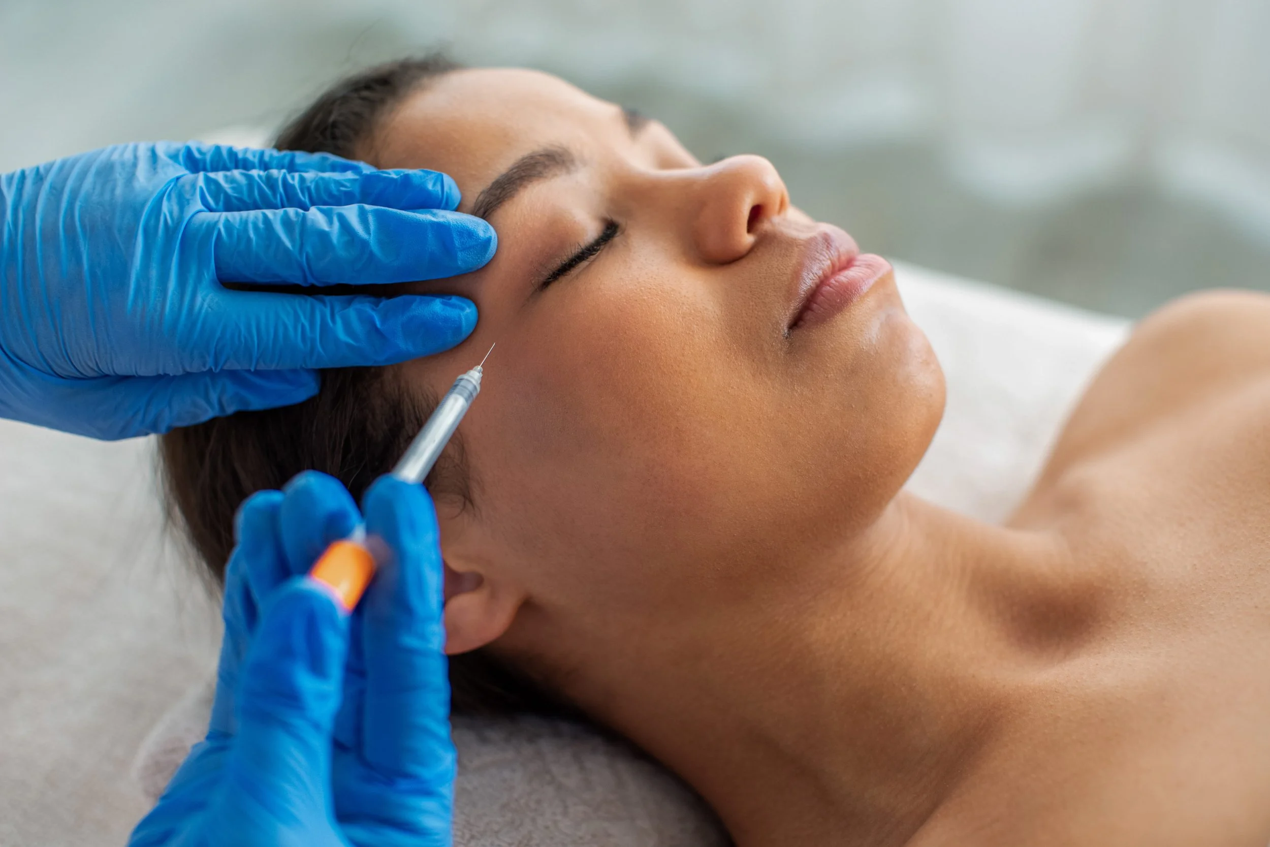 A woman lying down with her eyes closed, receiving an injection in her face from a healthcare professional wearing blue gloves.