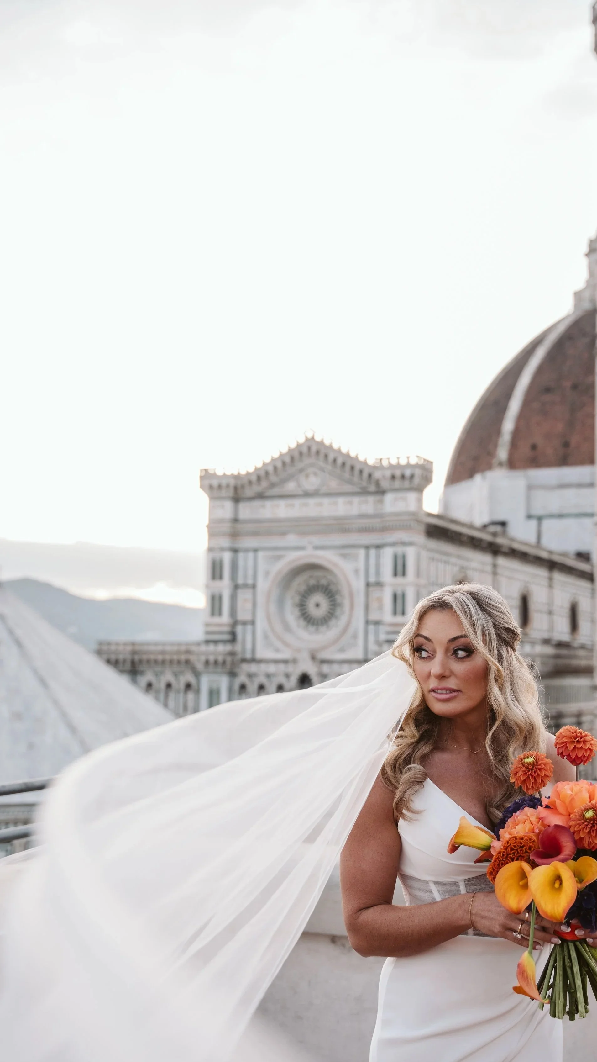 Una donna con un abito bianco tiene un bouquet di fiori arancioni, con il Duomo di Firenze sullo sfondo.