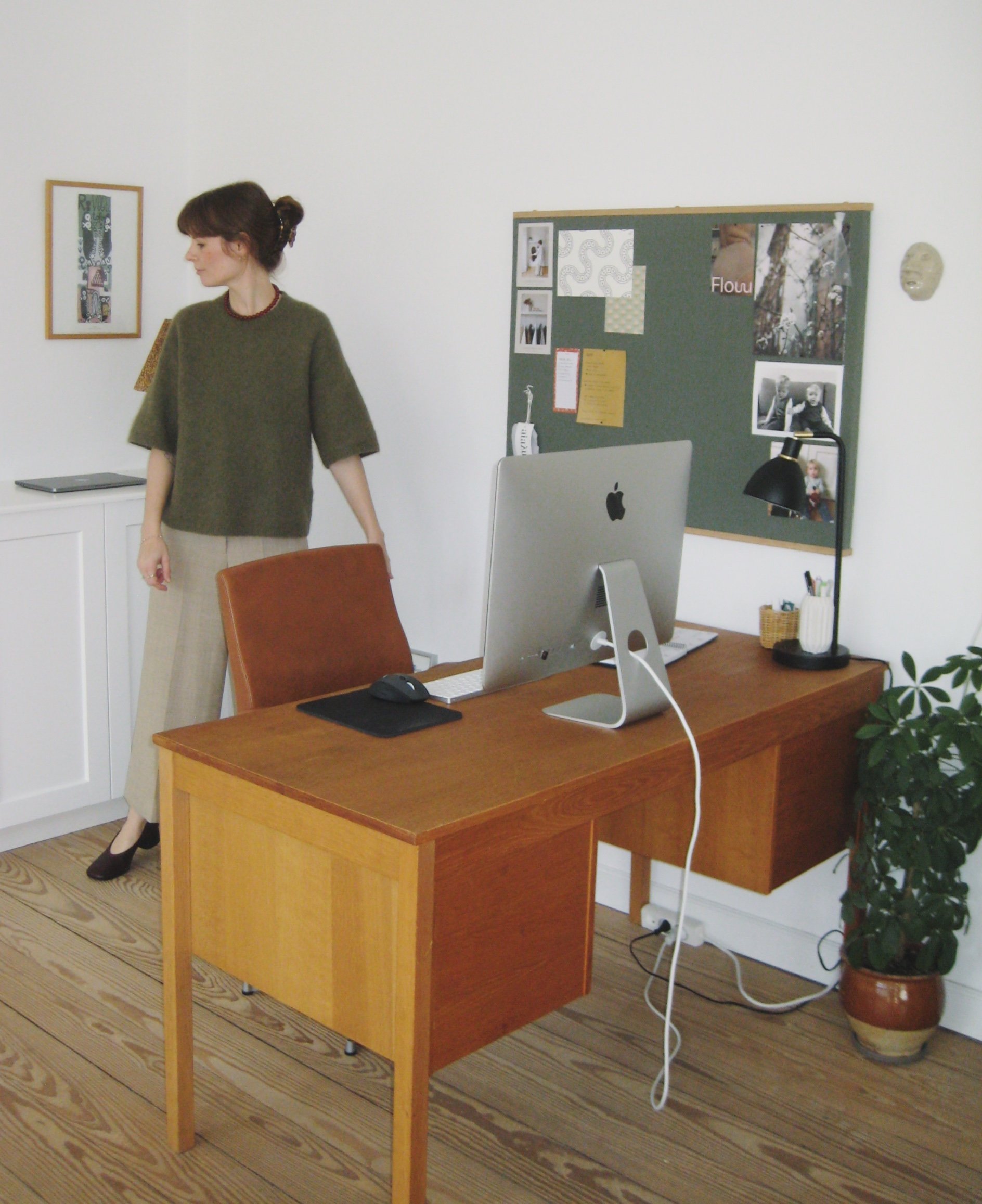 En kvinde står ved et kontorbord med en iMac-computer, der har en lamp ved siden af. Bag hende er der en opslagstavle med billeder og noter, og en plante i en potte. Der er et billede på væggen til venstre.