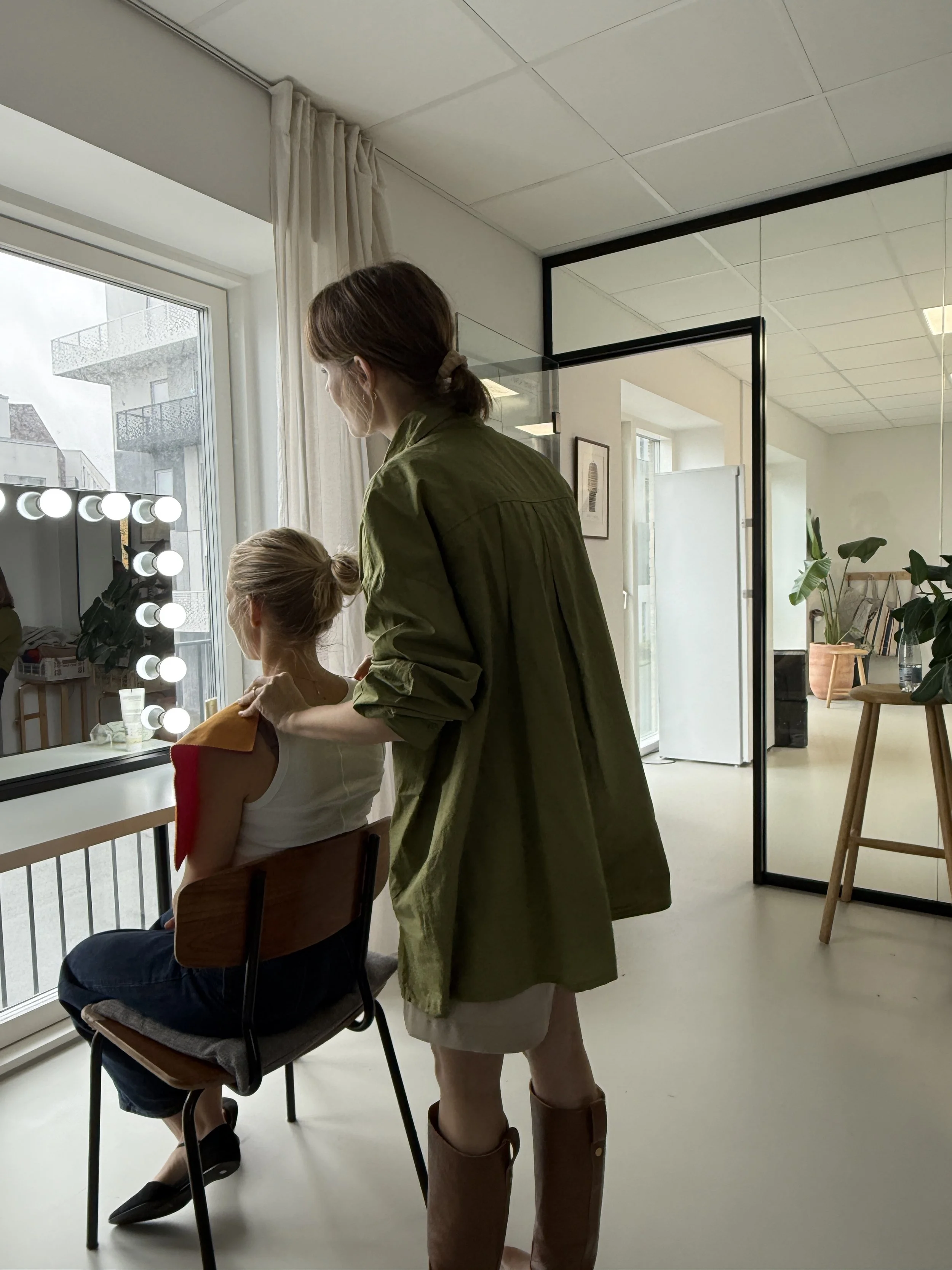En ung kvinde hjælper en anden ung kvinde med at få makeup på foran en spejl med lys rundt om. De sidder i et lysekrænt og moderne rum med store vinduer og grønne planter.