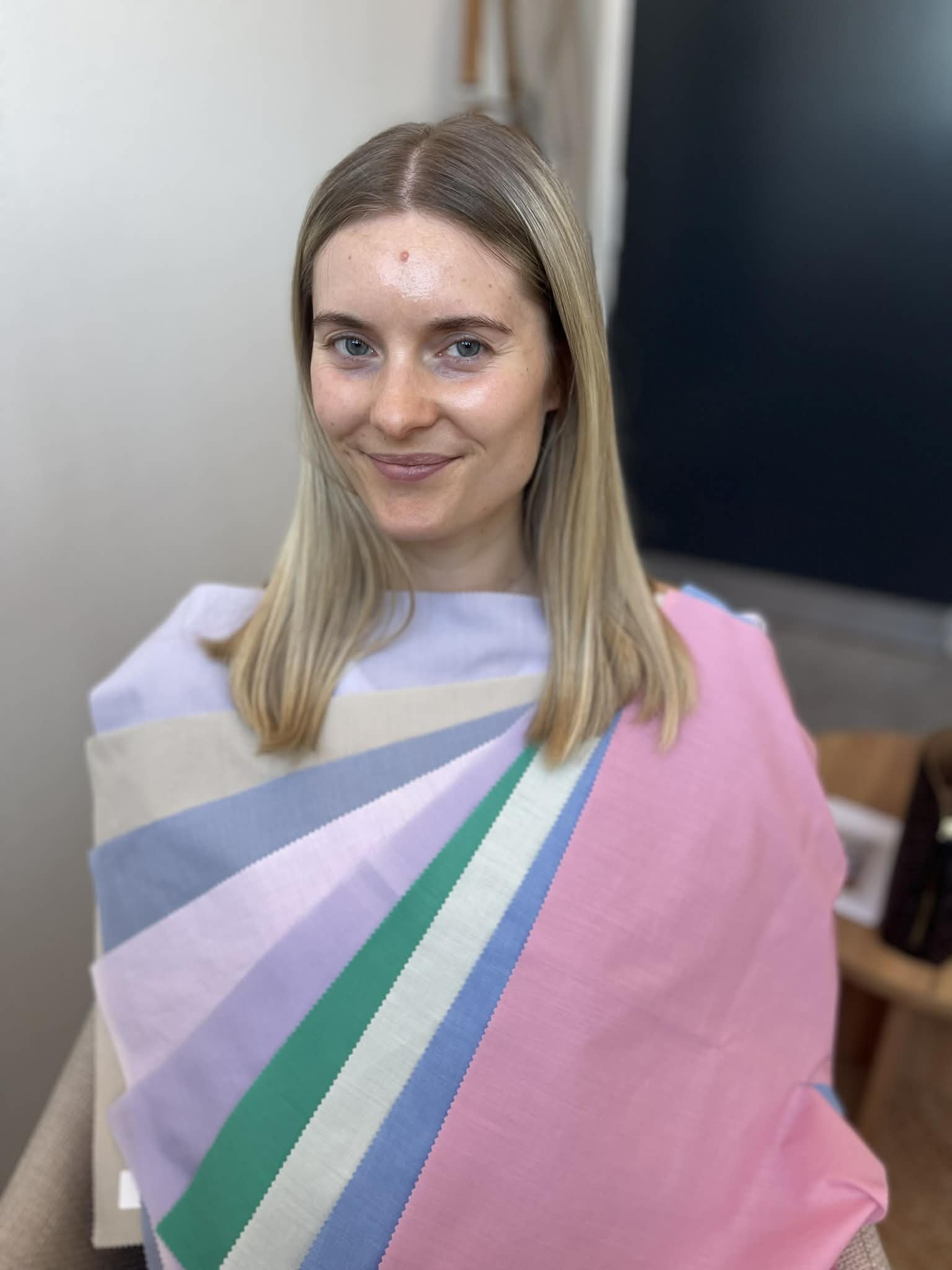En kvinde med blondt hår, der sidder i en stol, har noget på skuldrene, som er et farverigt stykke stof med striber i hvid, blå, grøn, lilla, og pink.