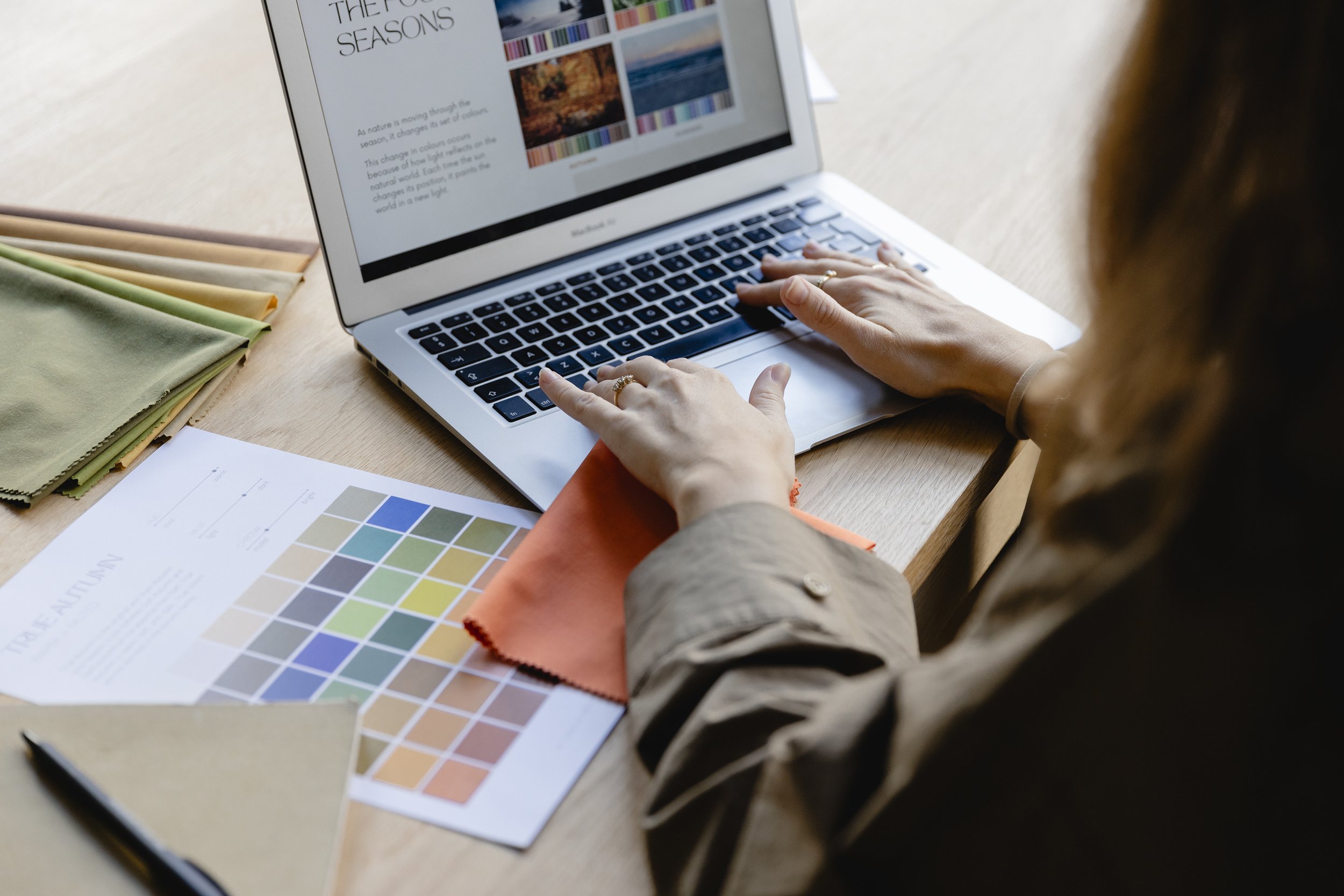 En person arbejder på en laptop, der viser en præsentation om de fire årstider. På bordet er der farvekort, papirer og stoffer.