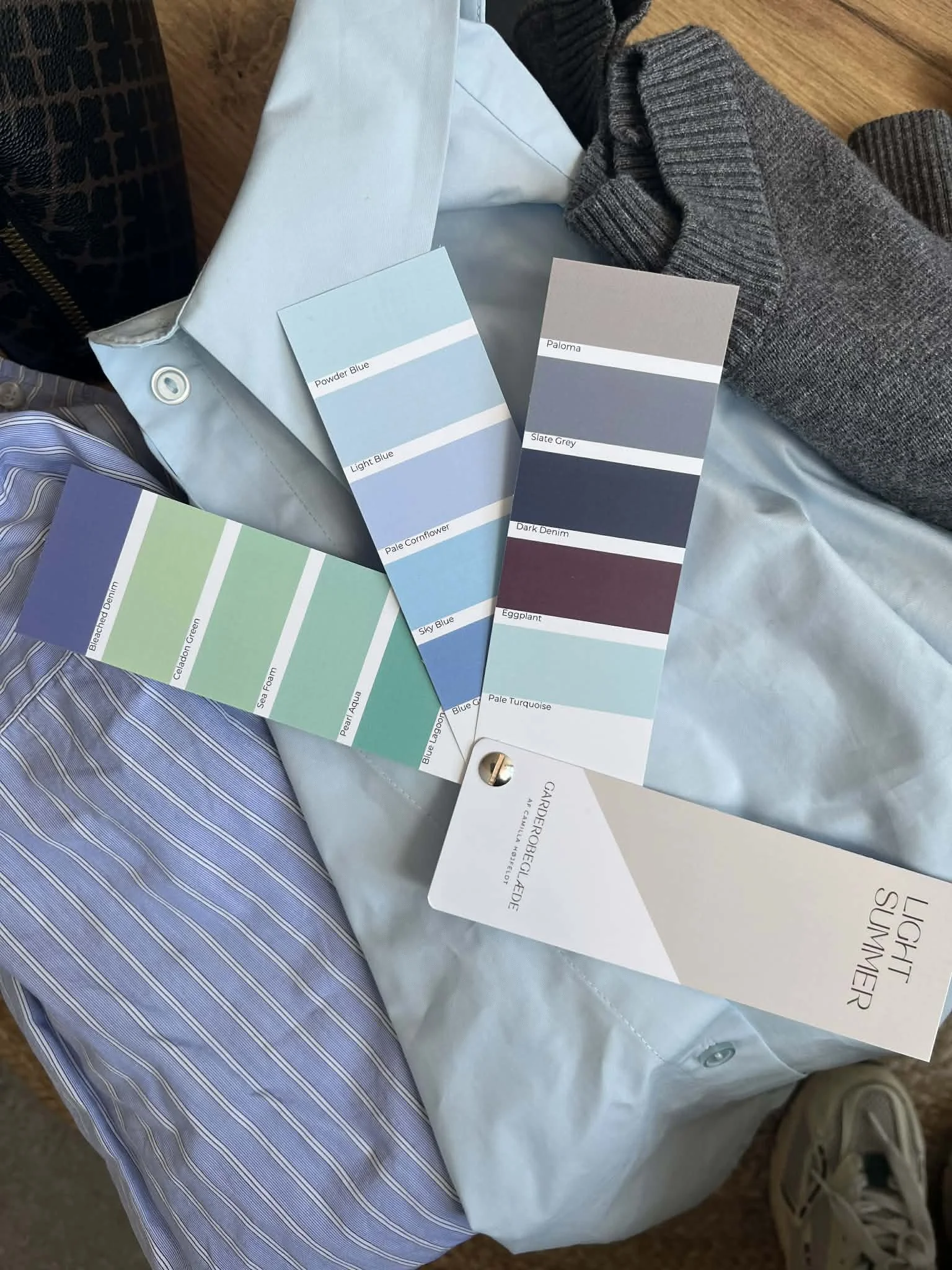 Billedet viser farvemøntprøver i nuancer af blå og grå, placeret på en pæn seng med tøj, herunder en lyseblå skjorte, en grå strikket sweater og en sko i den nederste højre hjørne.