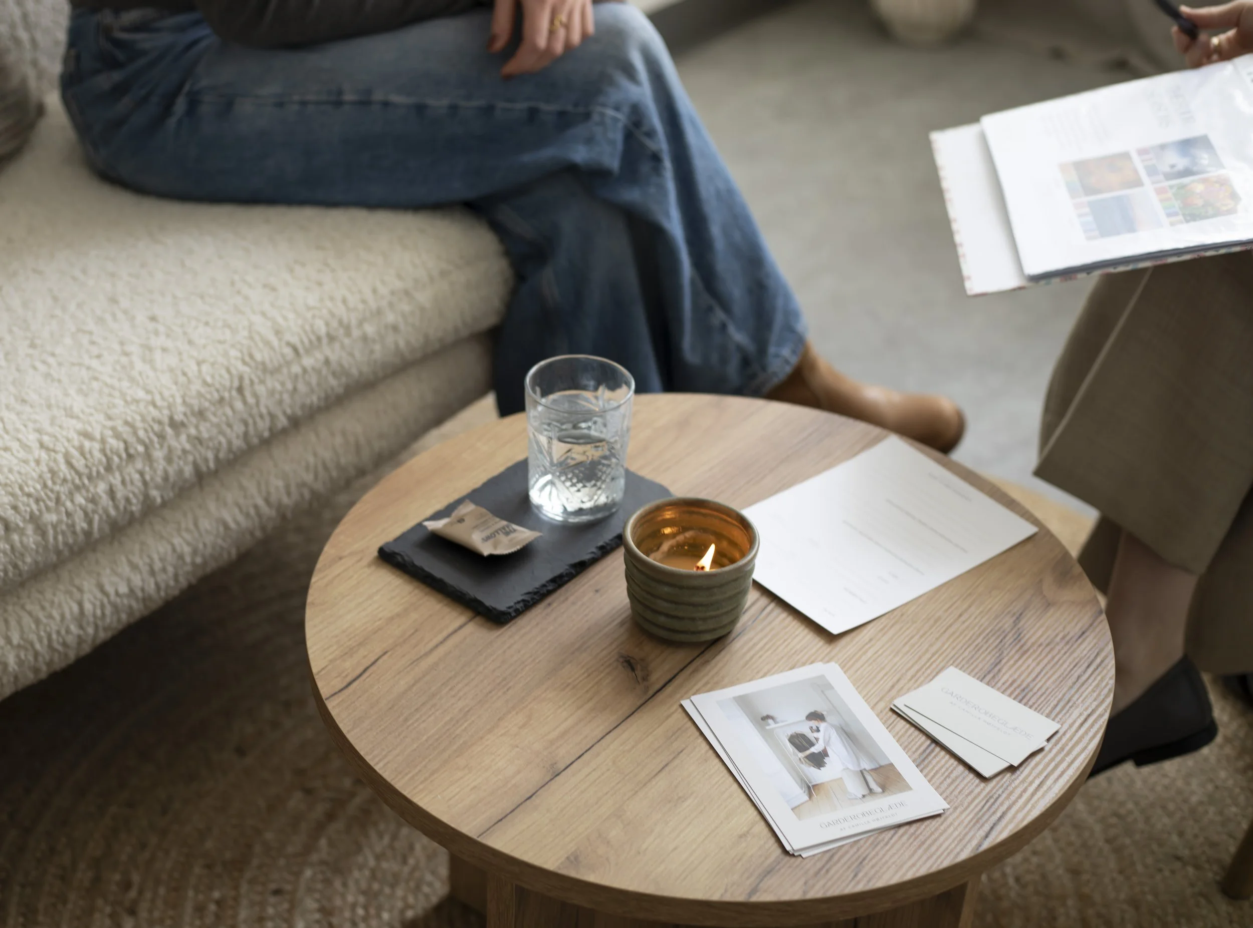 To personer sidder på en sofa, mens en anden person sidder ved et rundt træbord med brochurer og papir, der viser billeder af mennesker. Der er også et stearinlys, et glas vand og en kop på bordet.