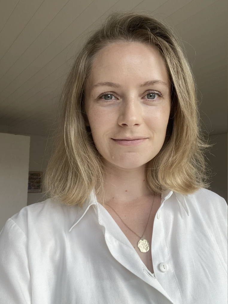 Et nærbillede af en smilende kvinde med mellemblond, skulderlangt hår, iført en hvid skjorte og guldhalskæde.